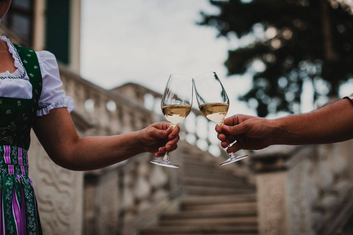 Zwei anstoßende Personen genießen Weißwein vor einem historischen Gebäude in Guntramsdorf, die Frau ist traditionell in Tracht gekleidet.