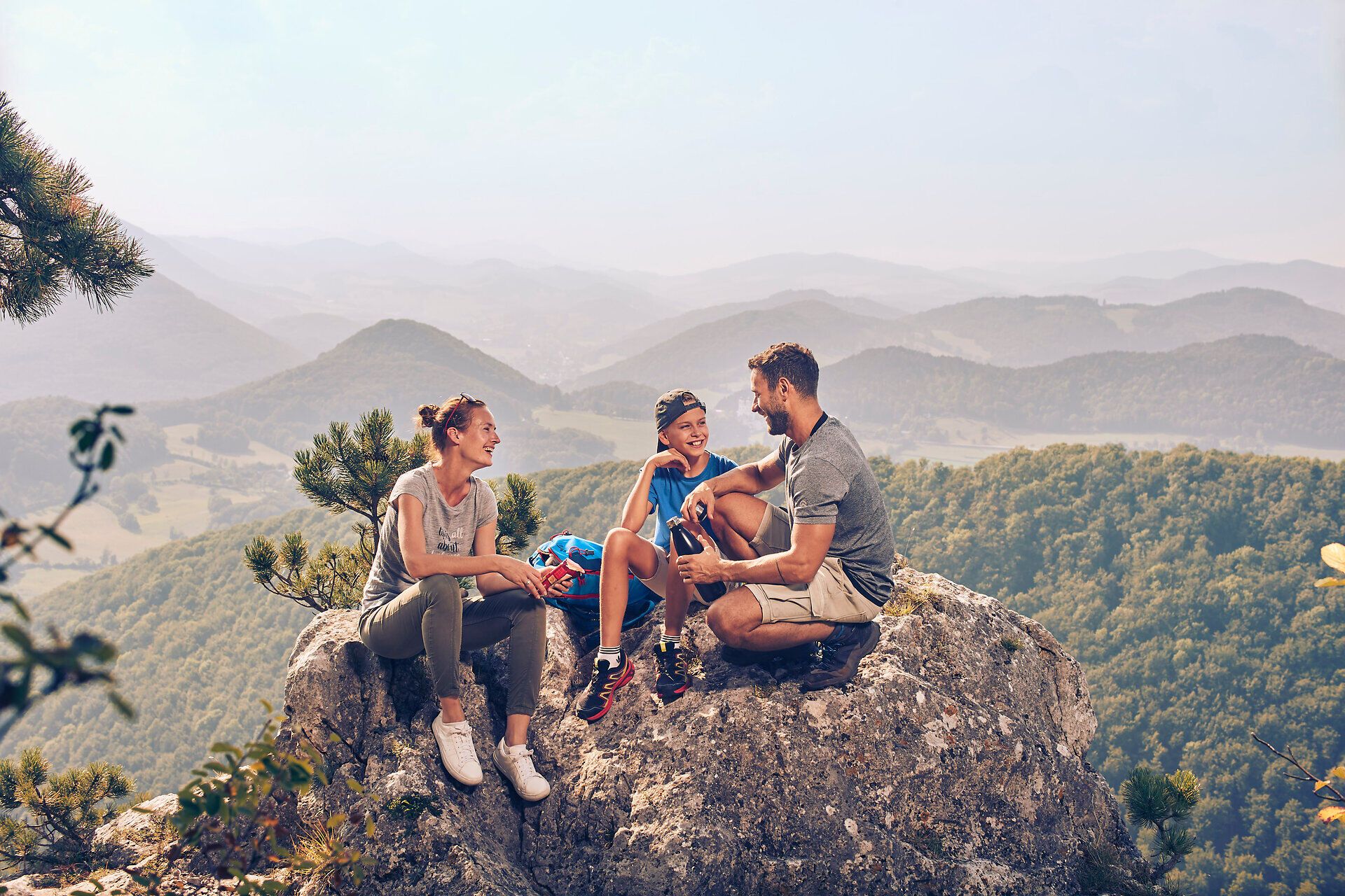 Eine Familie genießt eine wohlverdiente Pause auf einem Felsen mit atemberaubendem Blick auf die umliegenden Berge. Die frische Luft und die sanften Hügel laden zu weiteren Abenteuern in der Natur ein. Hier wird der Sommer in den Bergen zum unvergesslichen Erlebnis.