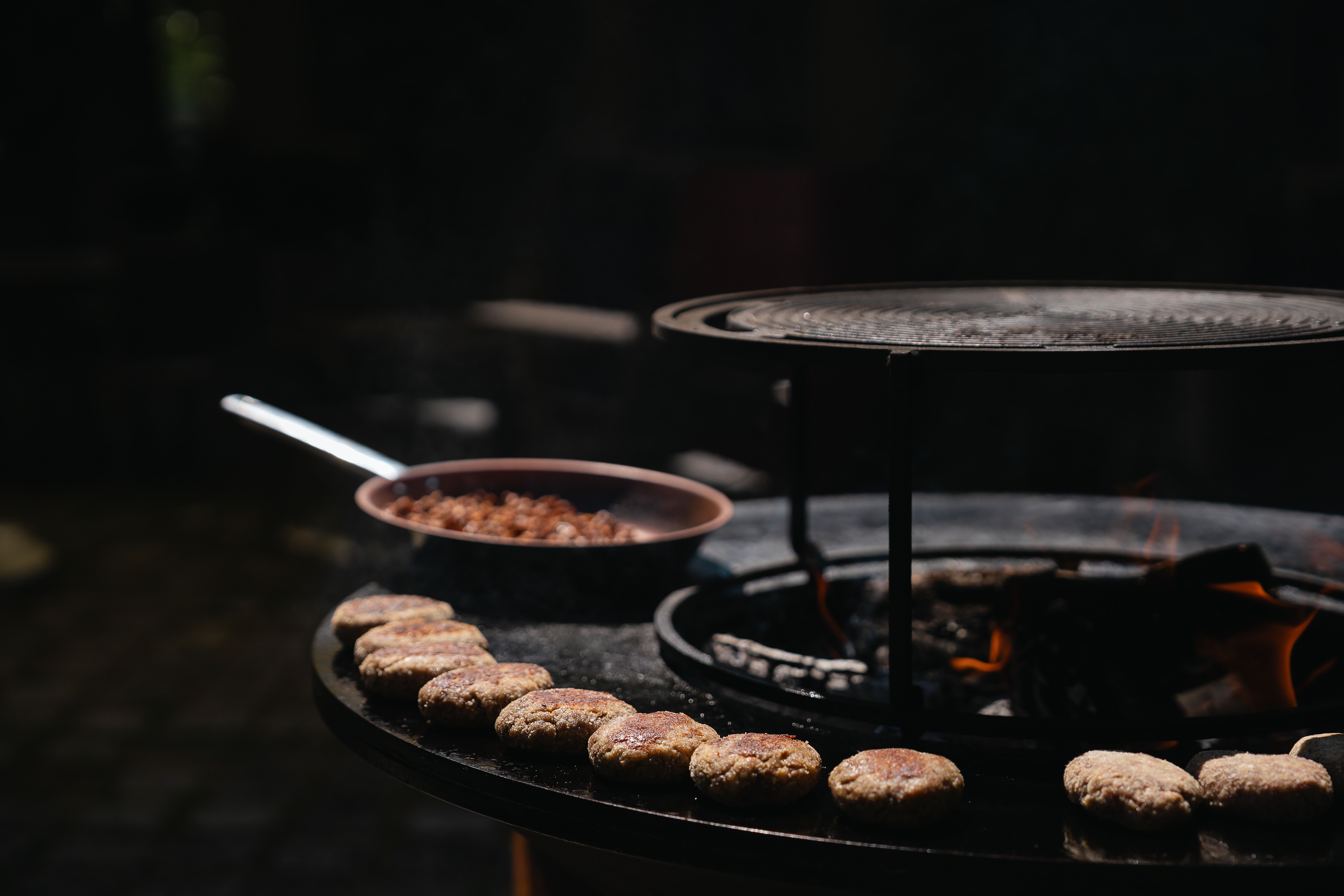 Hausgemachte Fleischlaibchen auf einer Feuerplatte beim Outdoor-Grillen, im Hintergrund eine Pfanne mit Zwiebelröster.