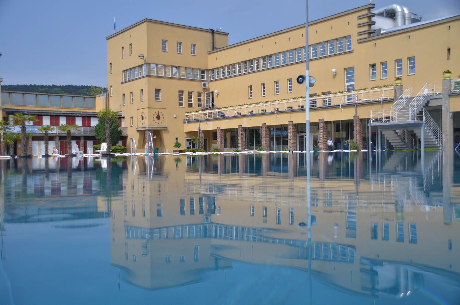 Ein Freibad mit einem großen gelben Gebäude im Hintergrund, das sich im Wasser spiegelt.