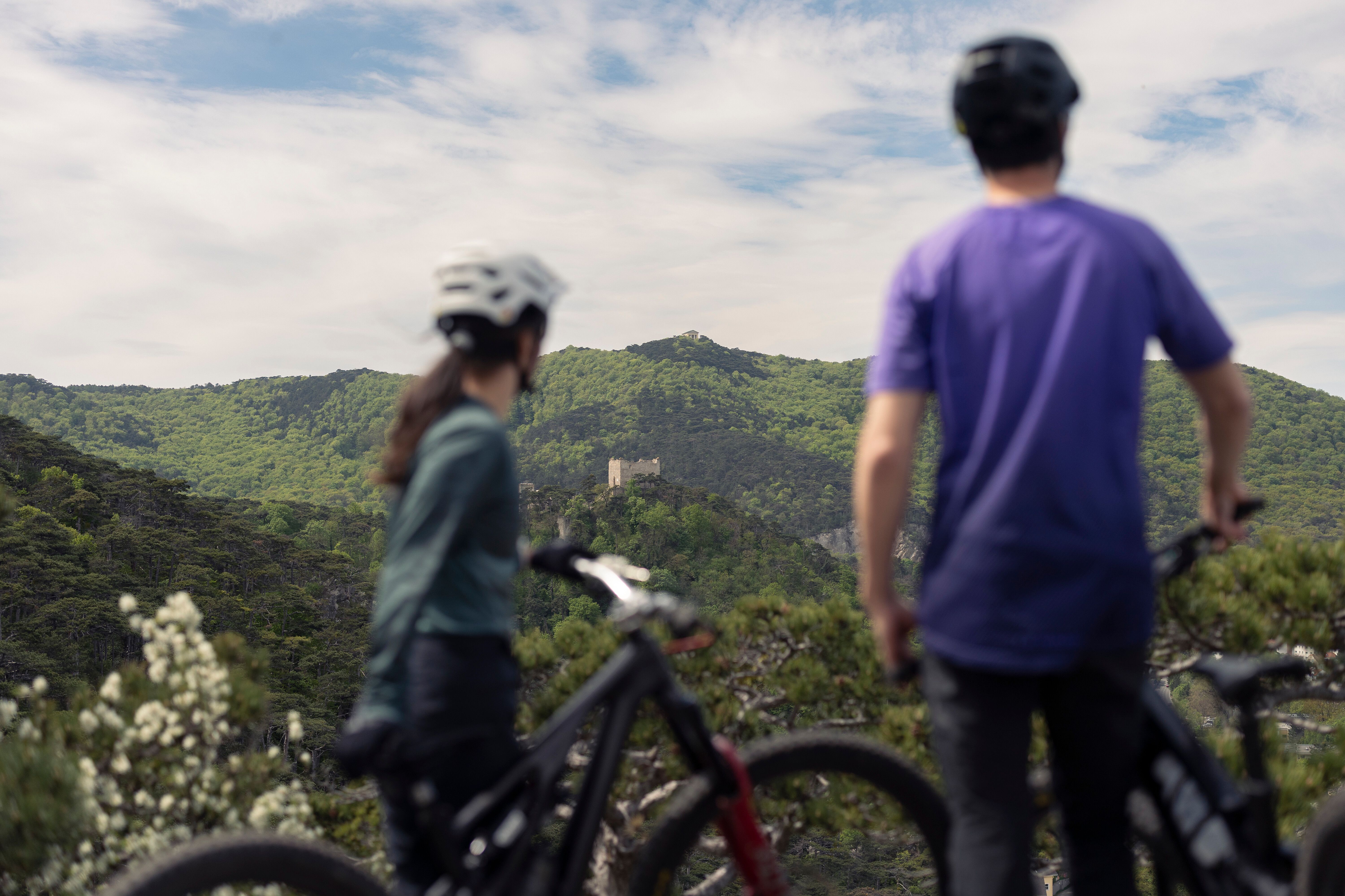 Eine Mountainbikerin und ein Mountainbiker stehen mit ihren Rädern und blicken über den dichten Wienerwald auf einen gegenüberliegenden Anninger, auf dem der Husarentempel thront – ein markanter Aussichtspunkt am Anninger südlich von Wien.