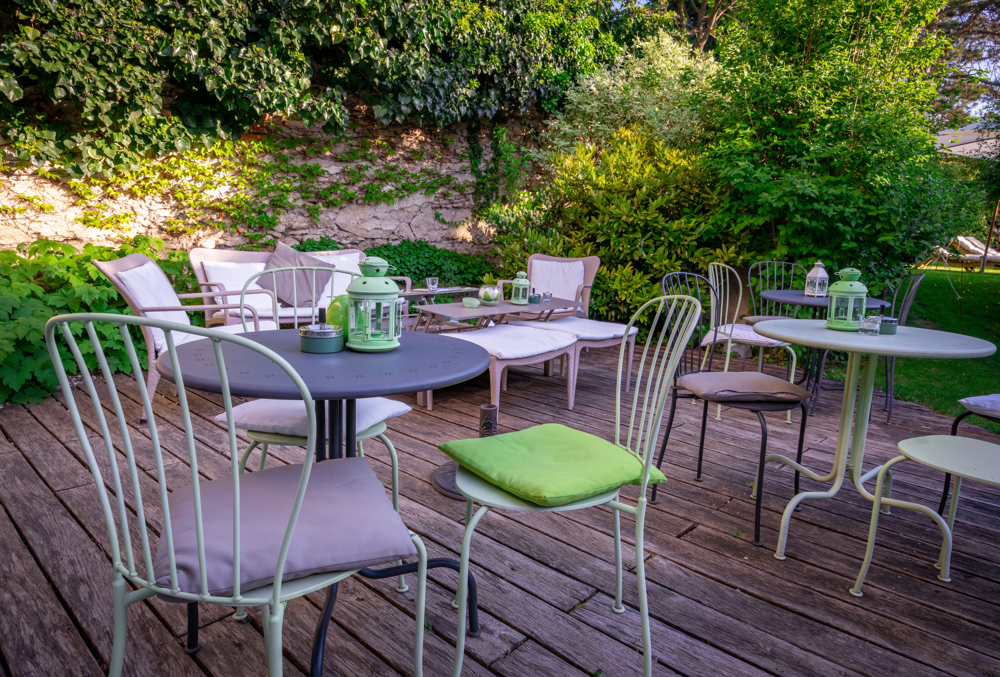 Gartenbereich mit Tischen und Stühlen auf Holzterrasse, umgeben von grünen Pflanzen.