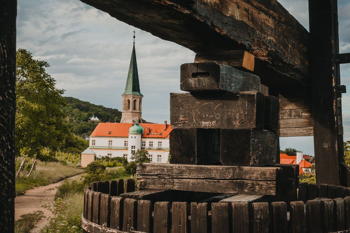 Eine traditionelle Weinpresse steht im Vordergrund, dahinter erstreckt sich ein Weingarten mit Schloss Gumpoldskirchen in sommerlicher Atmosphäre.