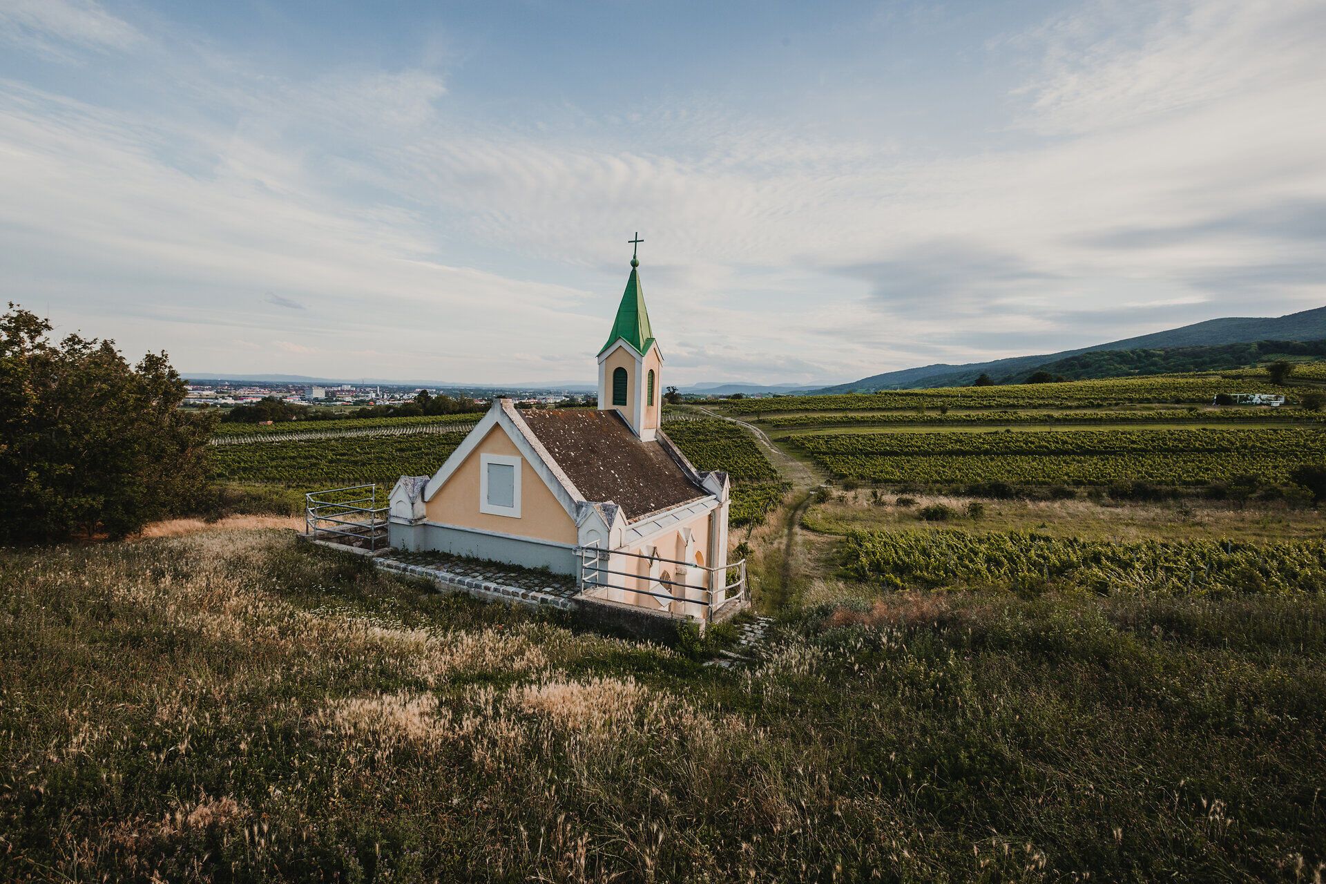 Umgeben von sanften Hügeln und üppigen Weinreben strahlt die kleine Kapelle eine friedliche Atmosphäre aus. Der grüne Turm und die pastellfarbenen Wände laden dazu ein, einen Moment der Ruhe und Besinnung zu genießen, während die Natur in voller Blüte steht.