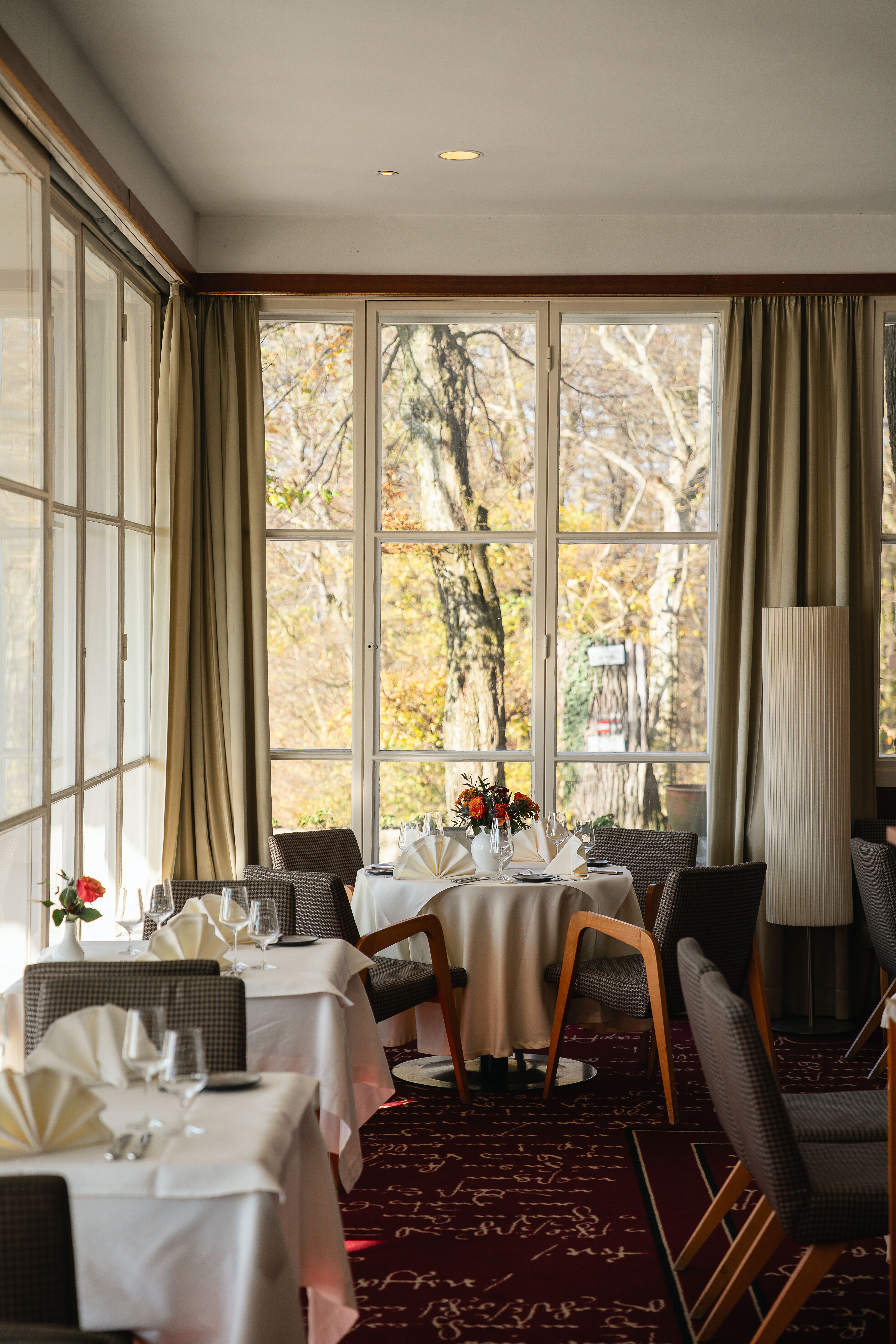 Ein elegant gedeckter Restaurantbereich im Berghotel Tulbingerkogel. Der Raum wird von hellem Licht durchflutet und bietet einen malerischen Blick auf die herbstlichen Bäume draußen, welche in warmen Farben leuchten.