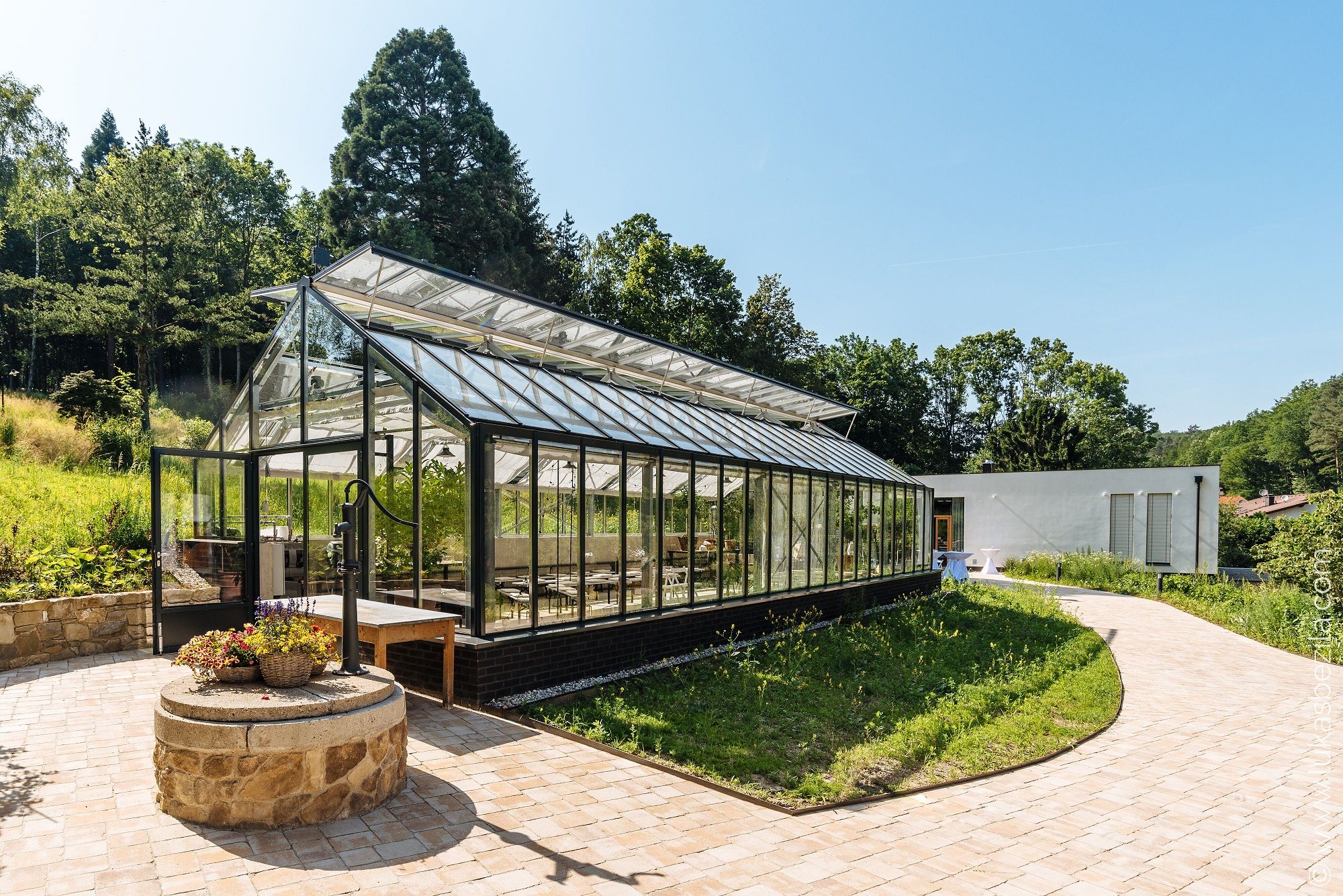 Ein modernes Gewächshaus mit Glaswänden und -dach steht in einer grünen Umgebung neben einem weißen Gebäude.
