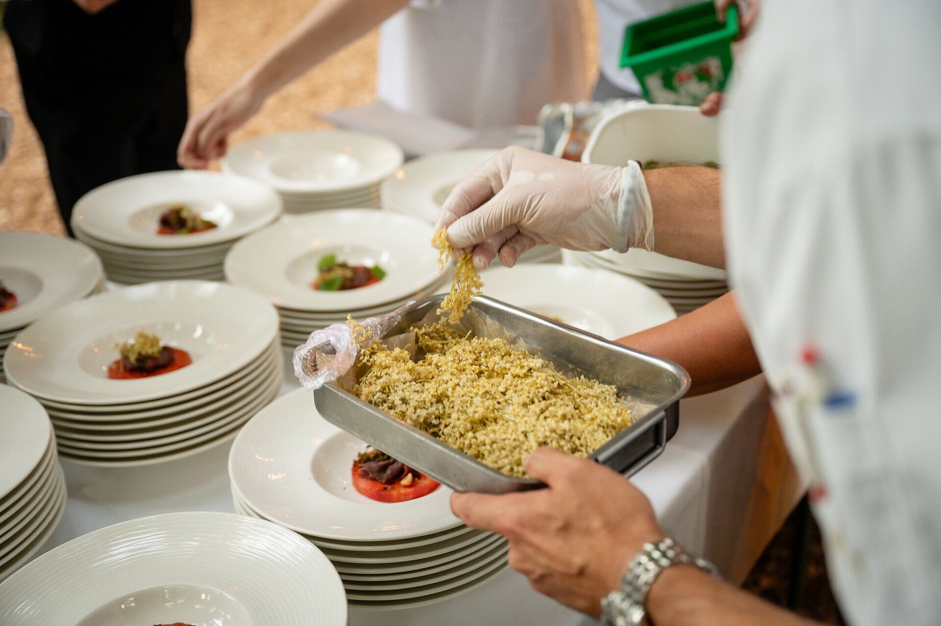 Auf einem Tisch beim Berghotel Tulbingerkogel werden weiße Teller liebevoll mit geräucherter Wassermelone, Haselnüssen und Hirschrohschinken für das Wienerwalddinner angerichtet. Holundervariationen fügen eine kunstvolle Dekoration hinzu, während die Szenerie des Wienerwalds in sanftem Sonnenlicht erstrahlt. Ein stiller Moment, in dem Natur und Kulinarik harmonisch verschmelzen.