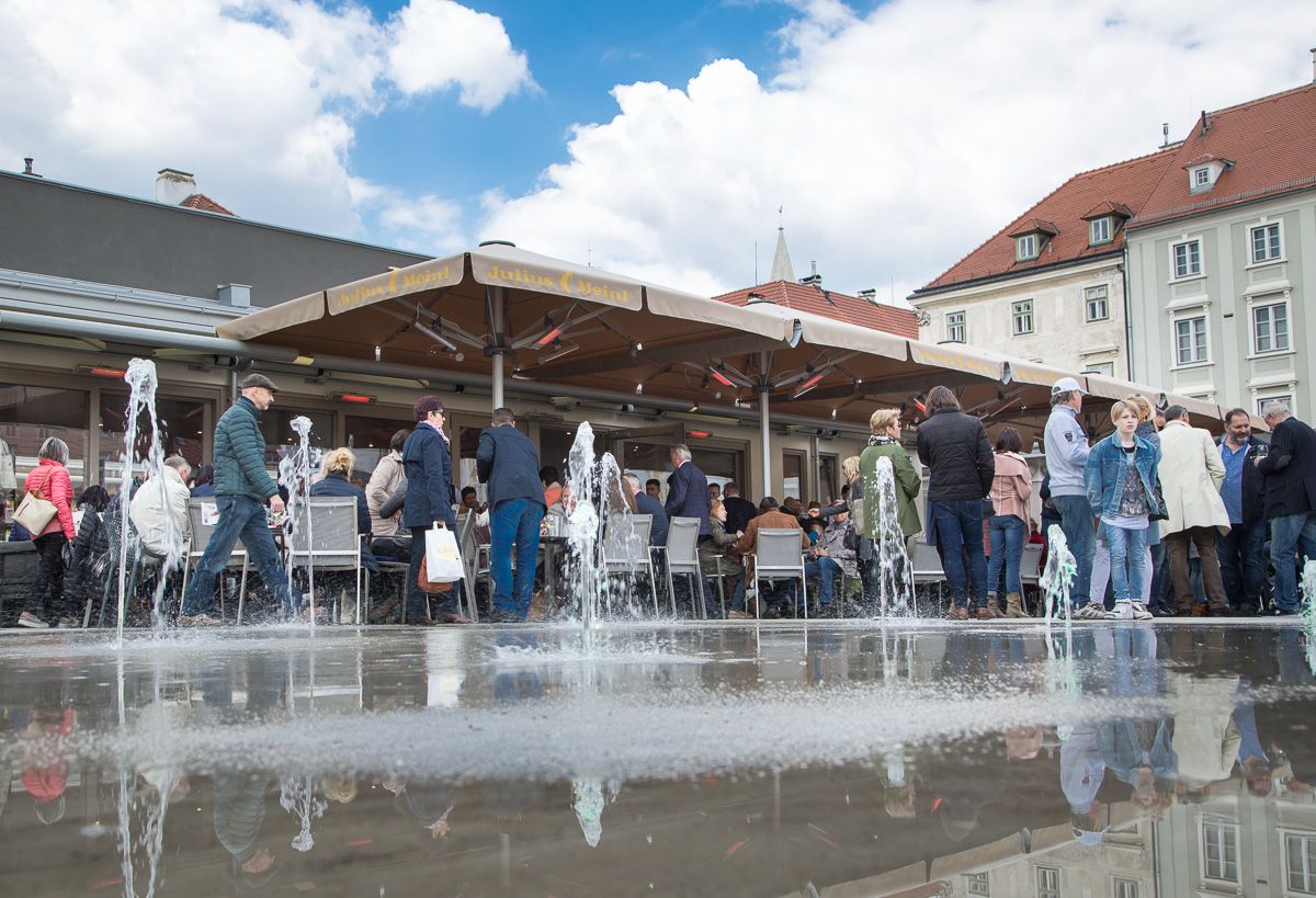 Menschen sitzen und stehen um einen Springbrunnen auf einem Platz mit Café.