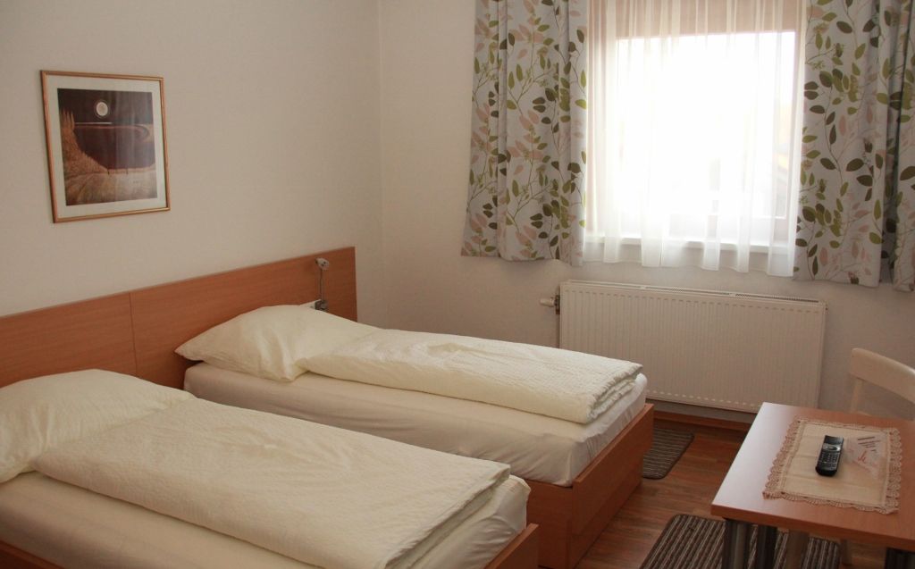 Ein Gästezimmer mit zwei Einzelbetten, einem Fenster mit Vorhängen, einem Tisch mit Fernbedienung und einem Bild an der Wand.