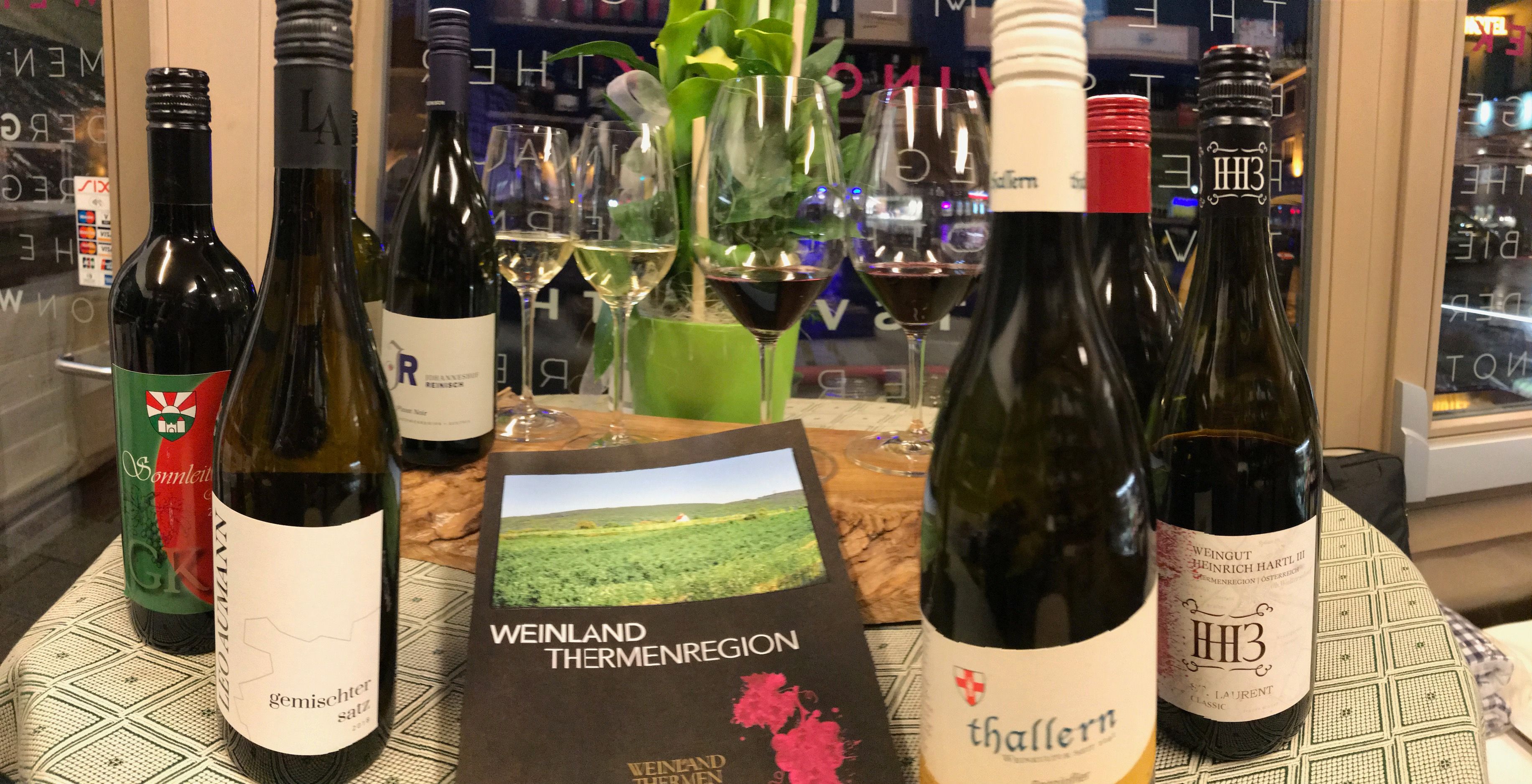 Weinflaschen und Gläser auf einem Tisch mit einem Prospekt der Weinland Thermenregion.