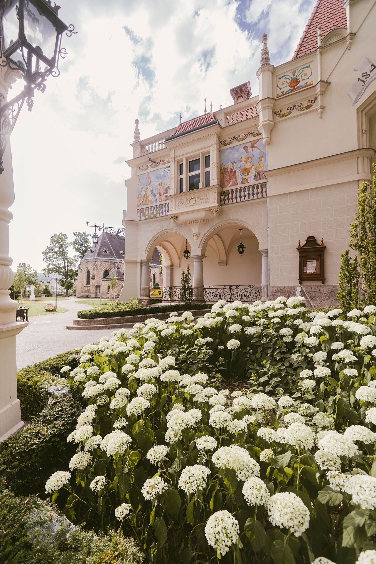 Umgeben von üppigen, blühenden Hortensien entfaltet sich die majestätische Fassade des Stadttheaters. Die kunstvollen Verzierungen und die harmonische Architektur laden dazu ein, in die kulturelle Vielfalt der Region einzutauchen. Ein Ort, an dem Kunst und Natur in perfekter Symbiose verschmelzen.