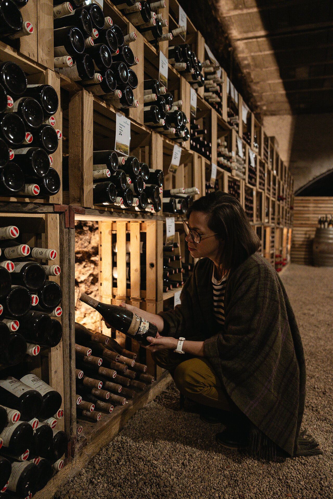 Katharina Graner kniet im Weinkeller des Freigut Thallern und betrachtet aufmerksam eine Weinflasche. Im Hintergrund erstreckt sich der weitläufige Weinkeller, der die Vielfalt des erlesenen Weinangebots präsentiert.