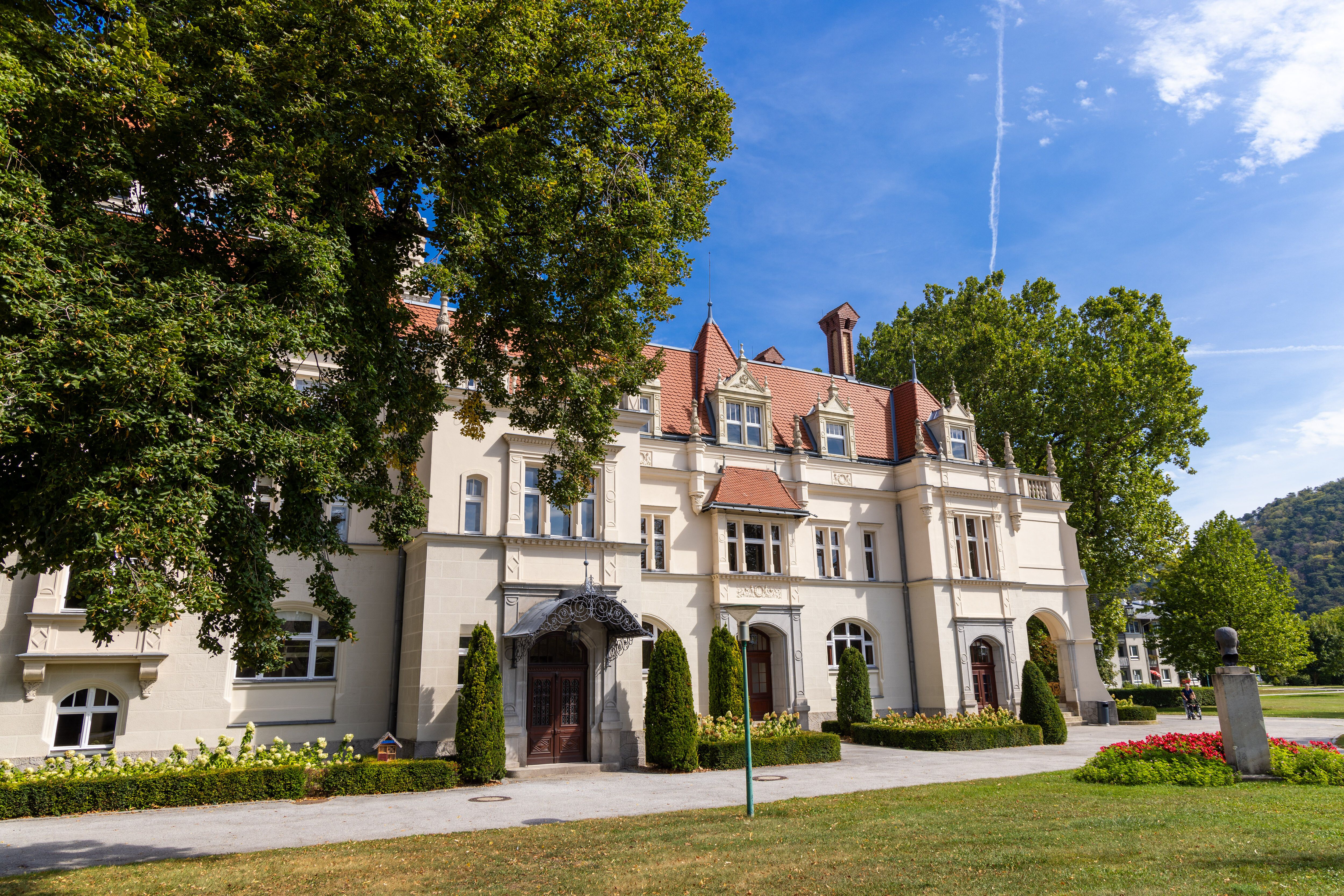 Das imposante Stadttheater Berndorf strahlt eine einladende Atmosphäre aus, umgeben von üppigem Grün und blühenden Blumen. Hier vereinen sich Kultur und Natur, während die sanften Hügel des Wienerwaldes im Hintergrund eine malerische Kulisse bieten.