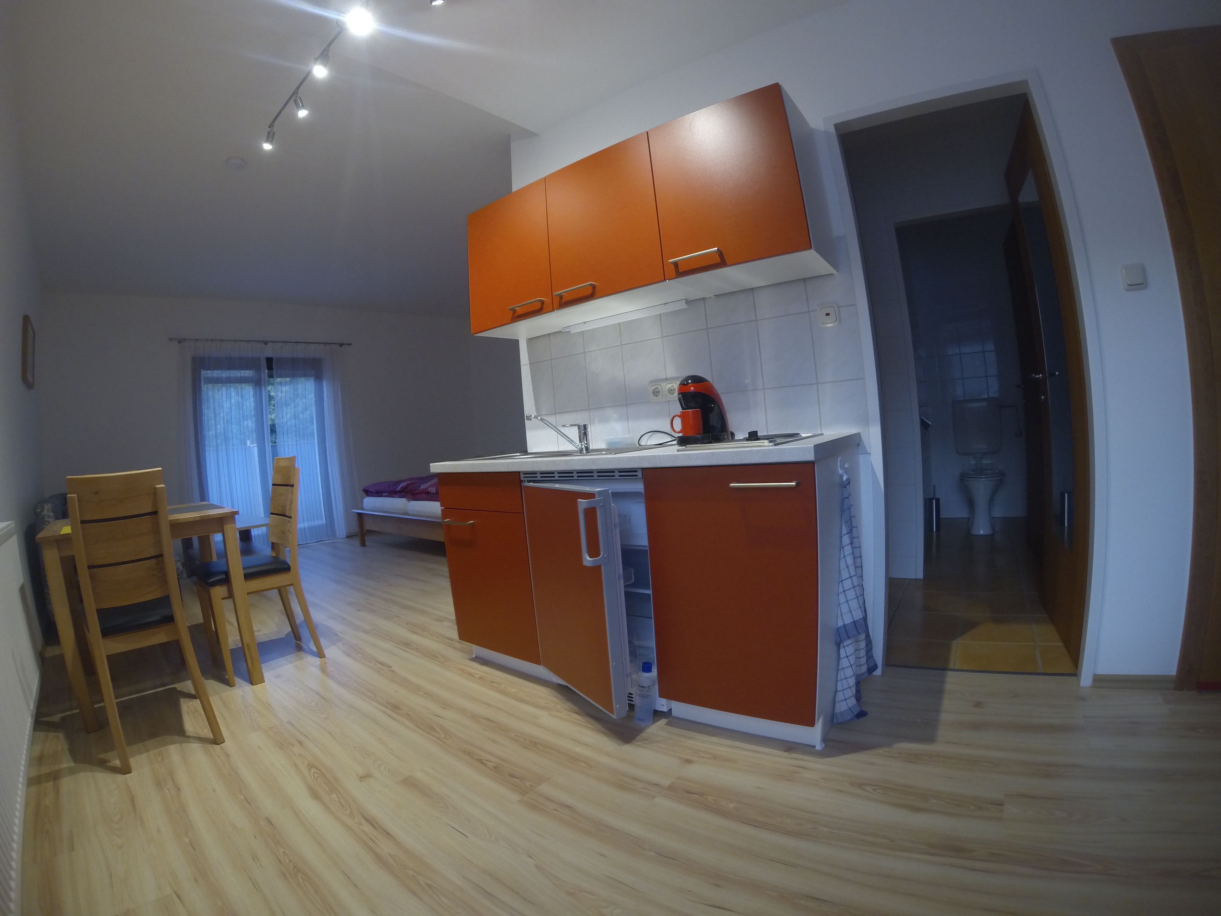 Kleine Küche in einem Apartment mit roten Schränken, Esstisch und Stühlen, Bett im Hintergrund und Badezimmer rechts.