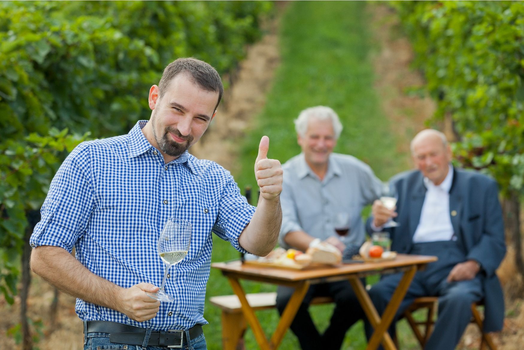 Ein Mann im karierten Hemd zeigt Daumen hoch und hält ein Weinglas, während zwei ältere Männer im Hintergrund an einem Tisch im Weinberg sitzen.