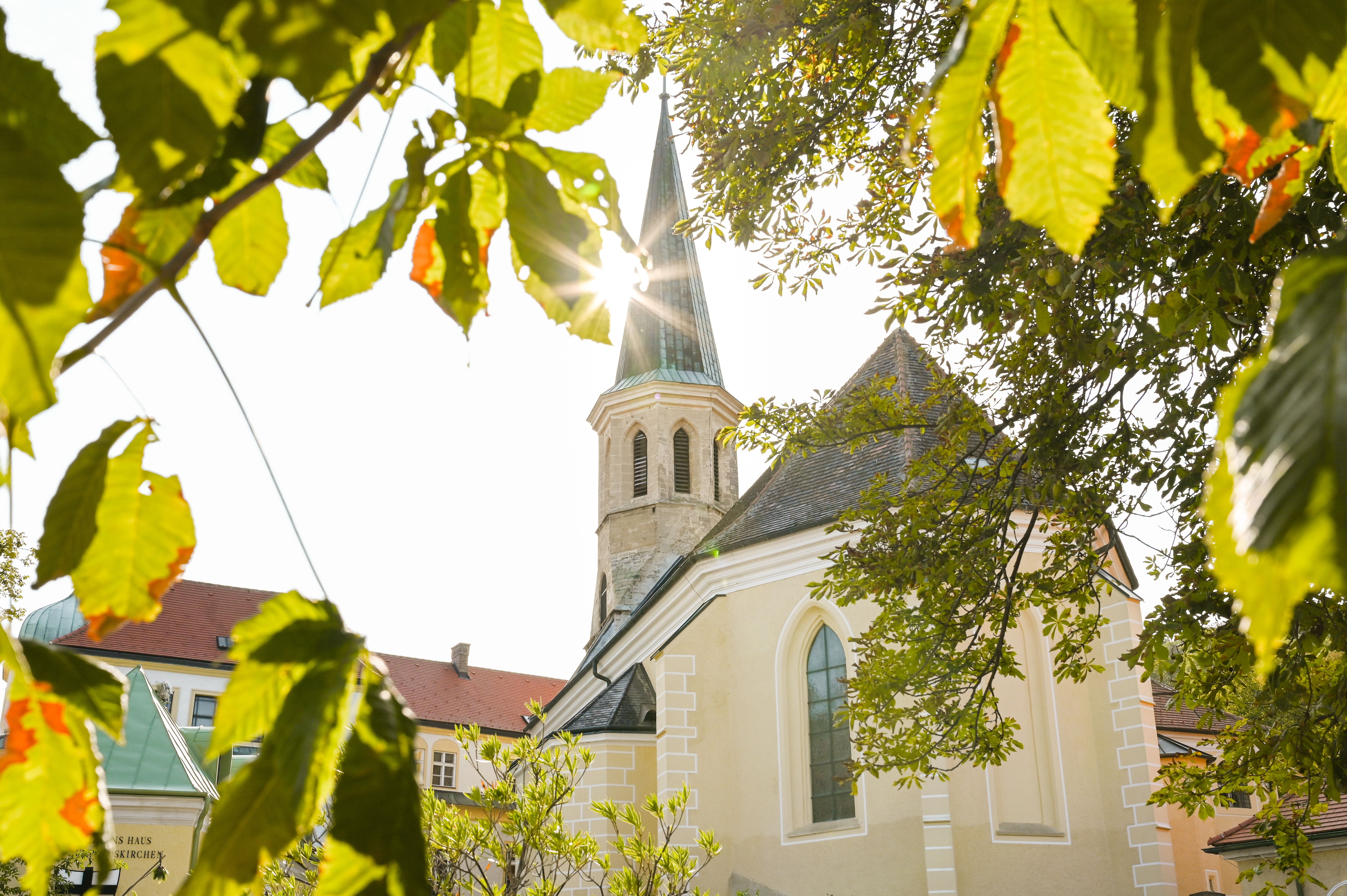 Die sanften Hügel umrahmen die charmante Kirche, deren Turm im Sonnenlicht strahlt. Umgeben von üppigem Grün und leuchtenden Blättern, lädt dieser Ort zum Verweilen und Entspannen ein. Hier spürt man die Ruhe und die Schönheit der Natur in vollen Zügen.