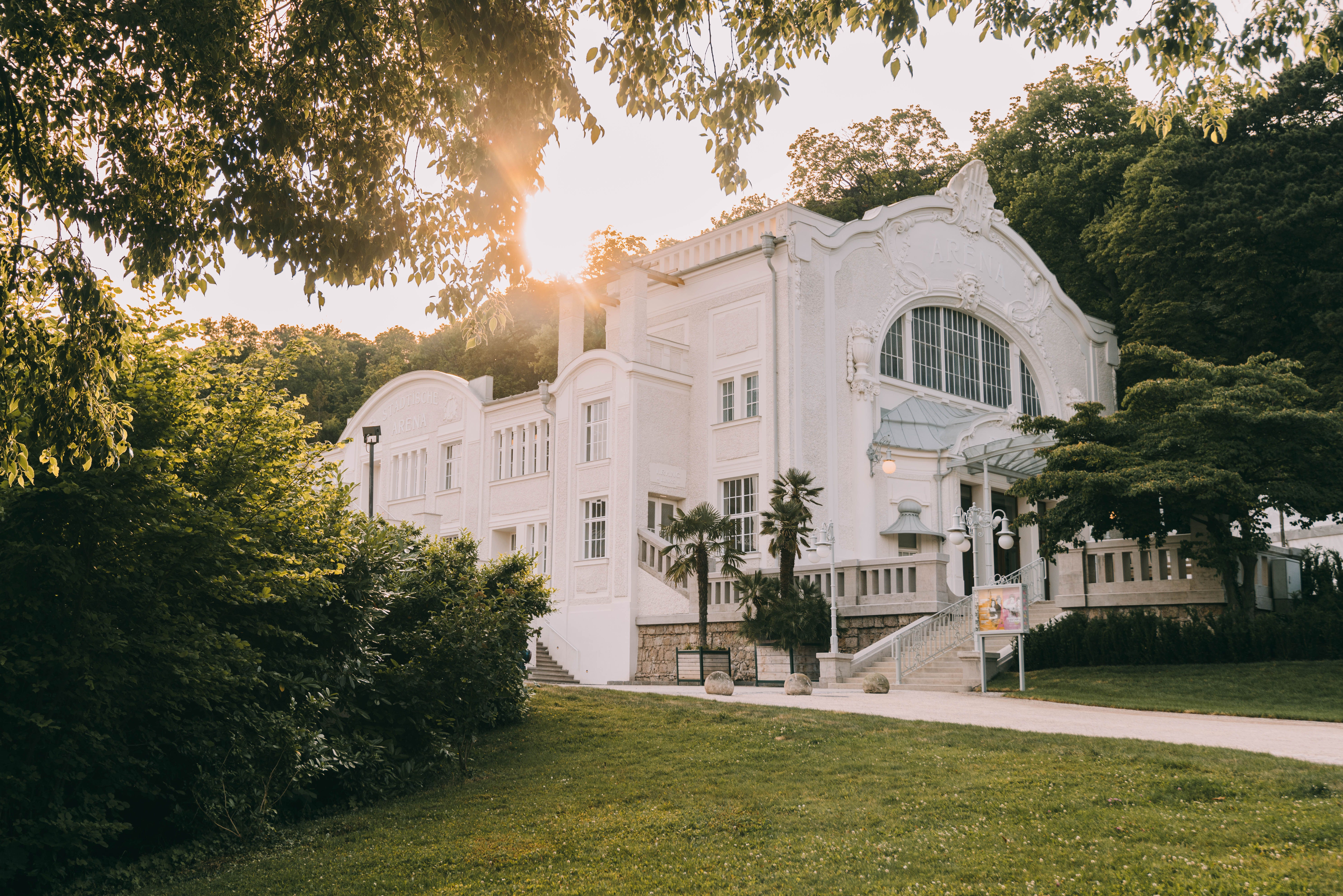Inmitten der malerischen Kulisse des Wienerwaldes strahlt das historische Gebäude in sanften Pastellfarben. Die warmen Sonnenstrahlen tauchen die Umgebung in ein goldenes Licht und laden dazu ein, die kulturellen Veranstaltungen des Theaterfests zu genießen. Üppige Pflanzen und gepflegte Grünflächen umrahmen diesen charmanten Ort und schaffen eine einladende Atmosphäre.
