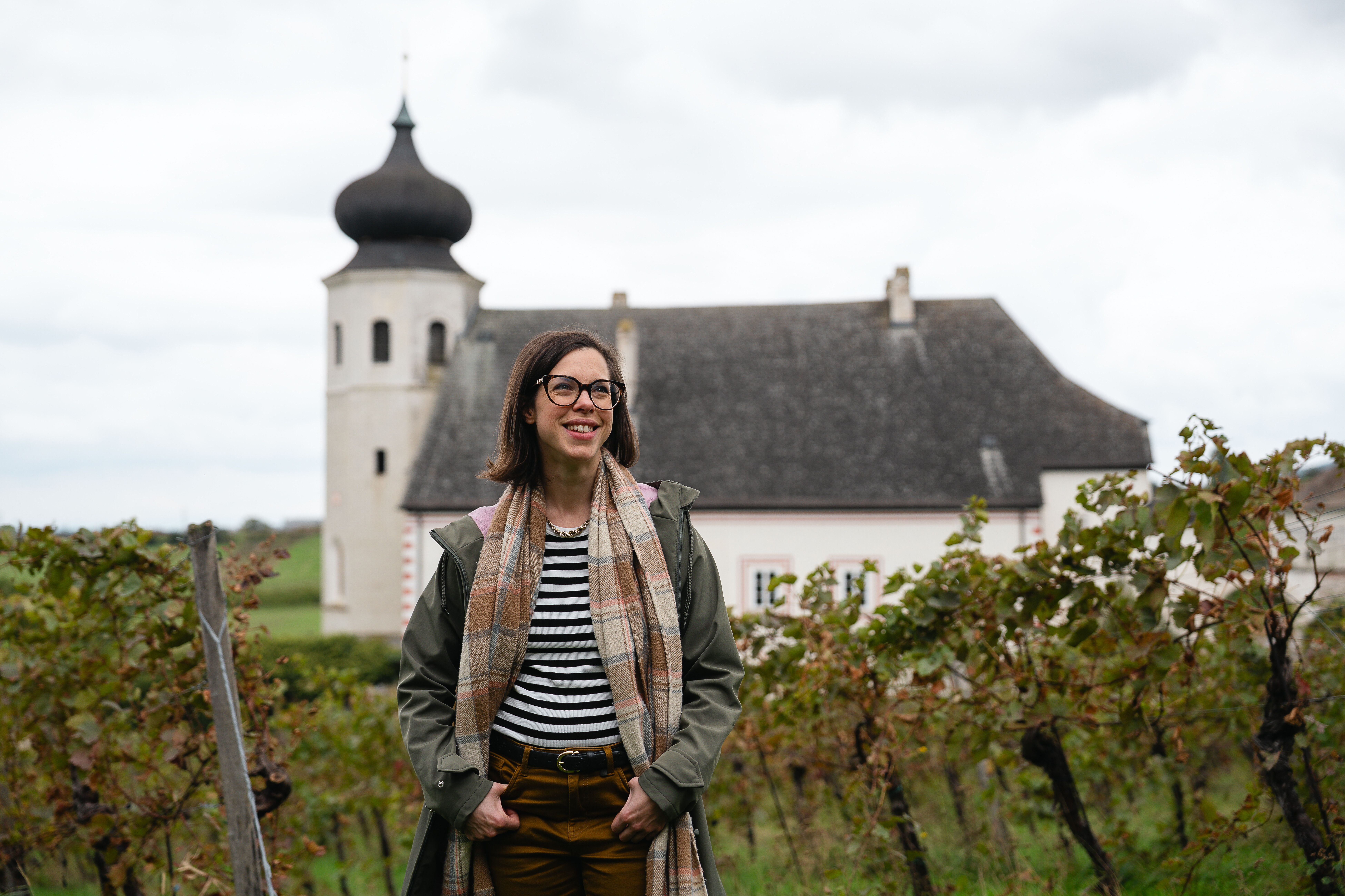Katharina Graner, die Geschäftsführerin des Freigut Thallern, steht lächelnd im Weingarten. Im Hintergrund ist das Weingartenhotel Thallern zu sehen, eingebettet in die reizvolle Landschaft der Weinberge.
