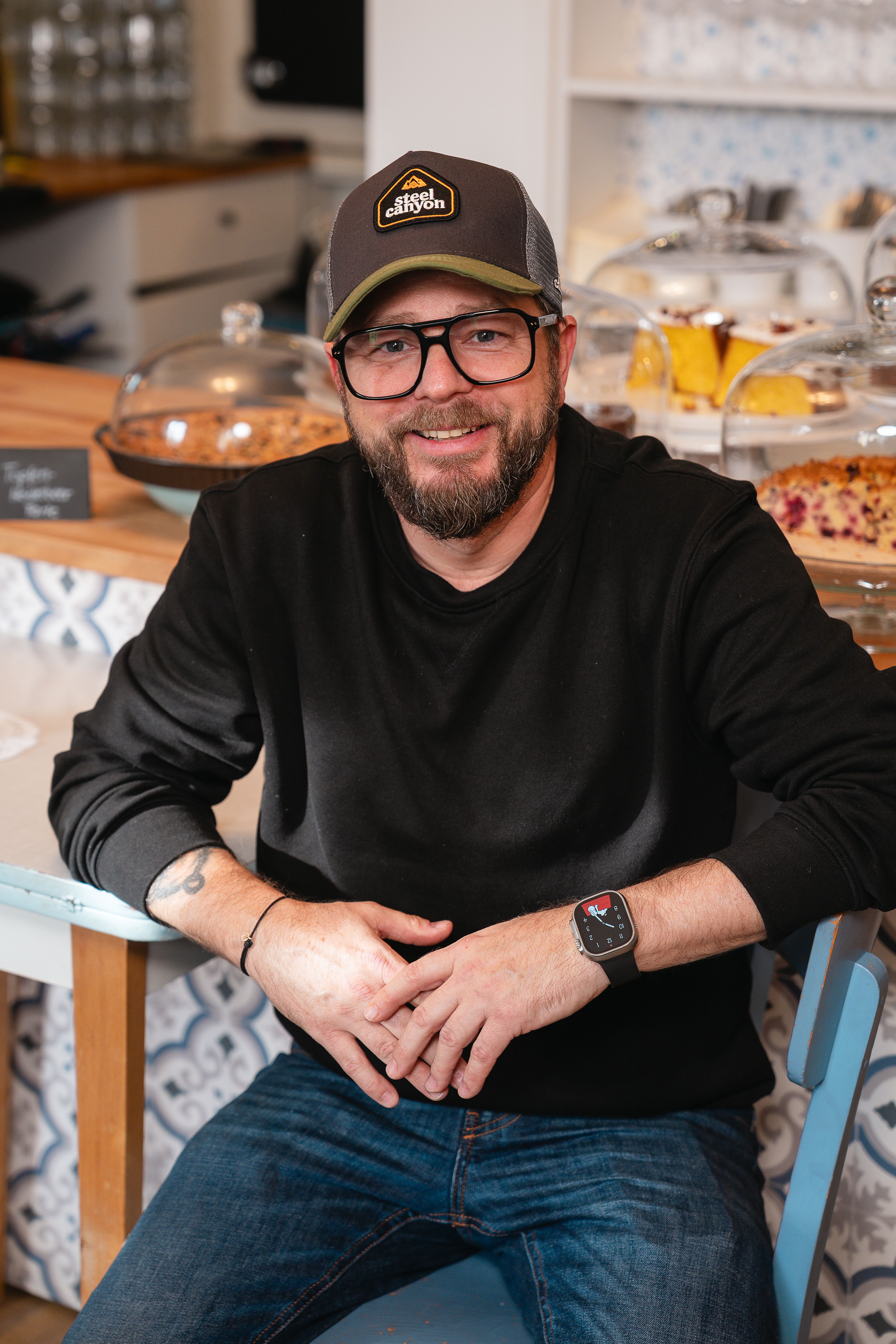 Michael Kulhanek sitzt entspannt in seinem Lokal "Kulhanek's Kaffee & Laden". Eine Reihe köstlicher Torten schmückt dahinter die Theke . Ein Ort, wo Genuss im Licht badet und die süßen Versuchungen voller Erwartung strahlen.