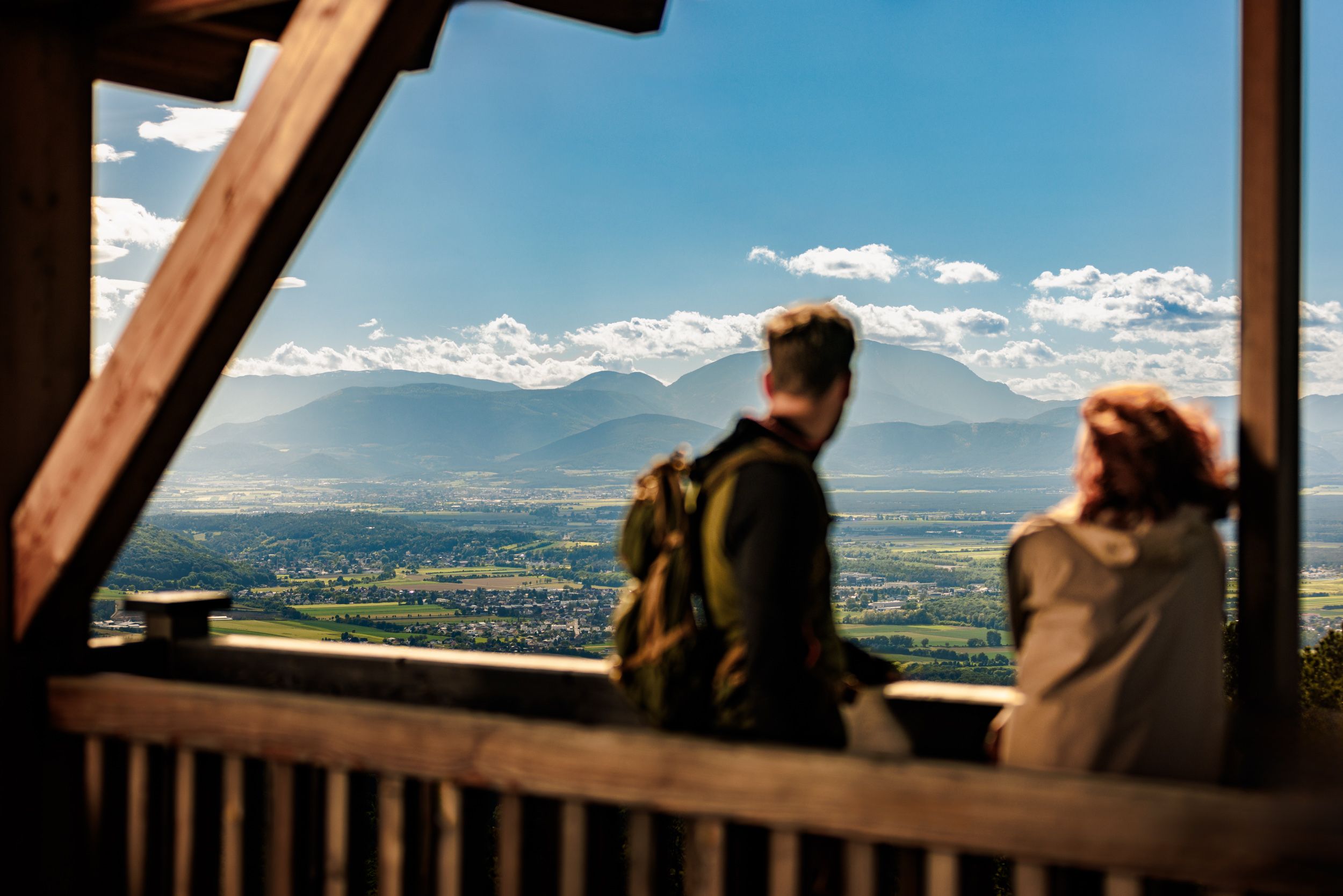 Zwei Personen stehen auf der Plattform des hölzernen Aussichtsturmes Lanzenkirchen/Wiesen, im Hintergrund ist eine weite Ebene und am Horizont der Schneeberg gut erkennbar.