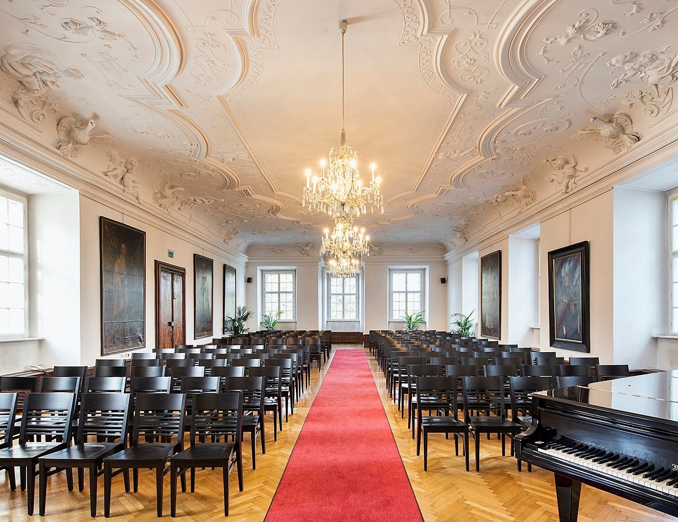 Elegant hall with stucco ceiling, chandeliers, rows of chairs and red carpet.