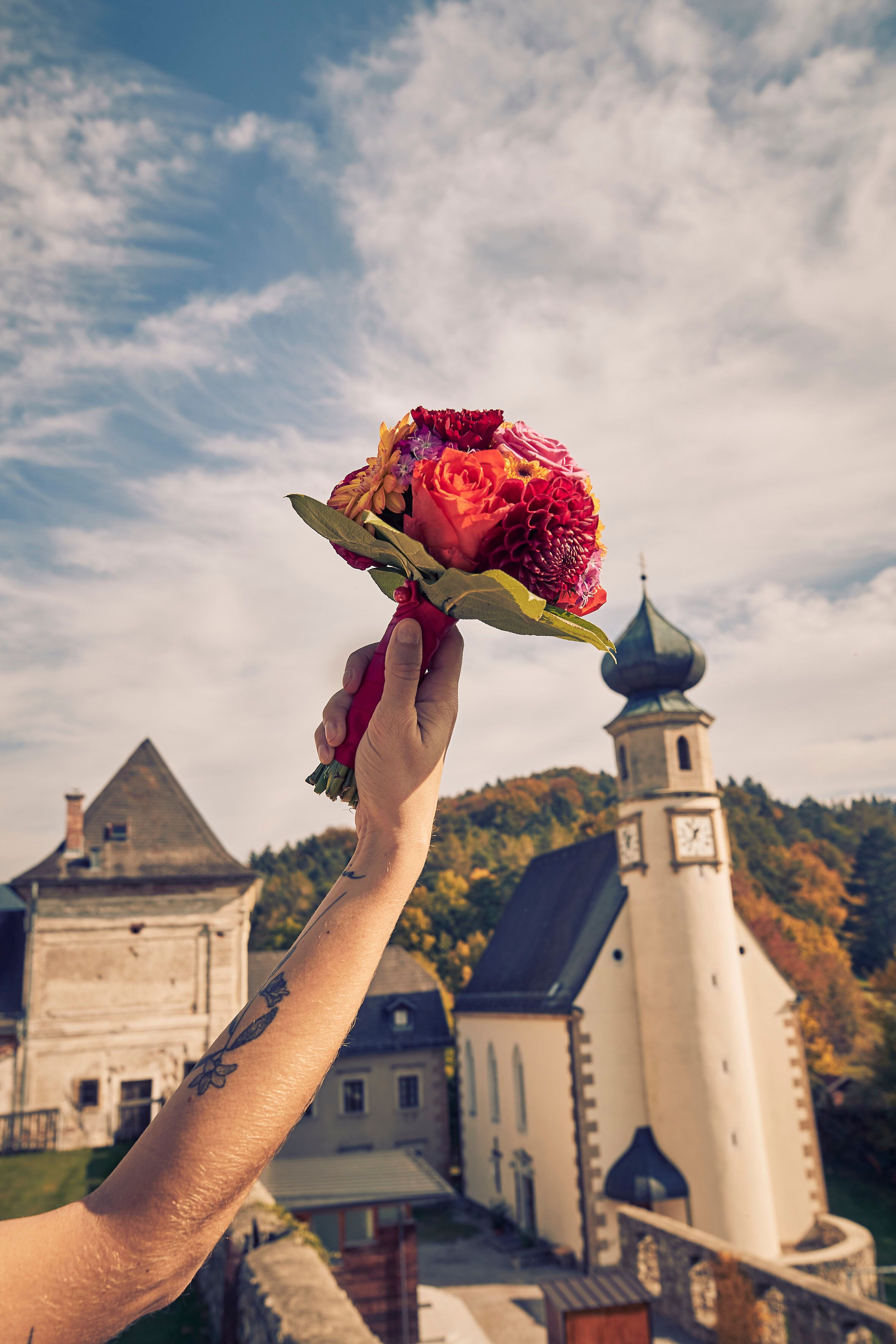 Ein strahlender Sommertag umhüllt die malerische Kulisse des Schlosses Hernstein, während ein bunter Blumenstrauß in der Hand gehalten wird. Die sanften Hügel und die historische Architektur schaffen eine romantische Atmosphäre, die zum Verweilen einlädt.