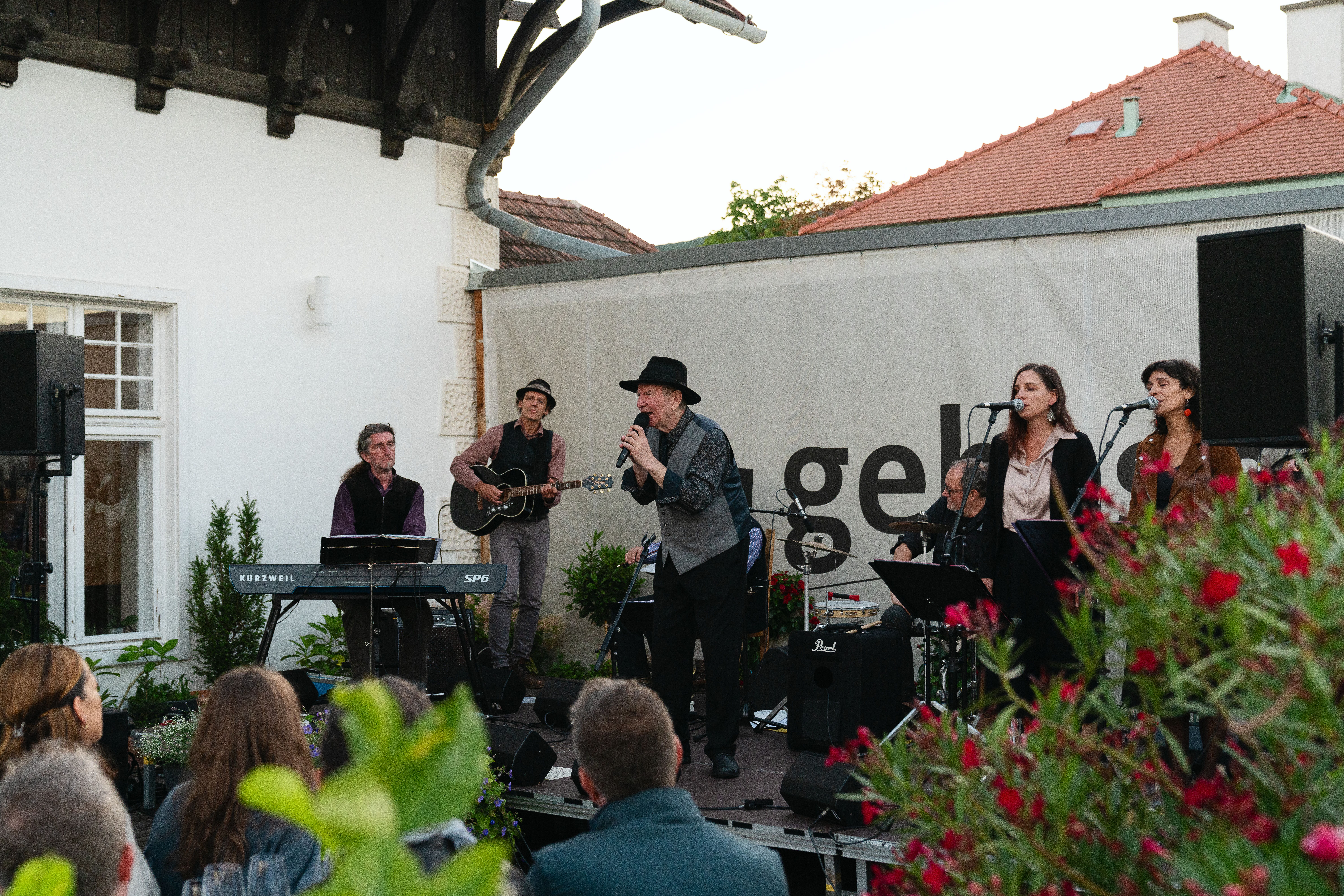 Ein lauer Sommerabend erfüllt die Luft mit Musik und Genuss. Die Klänge einer Tribute-Band, die Leonard Cohen huldigt, umgeben die Gäste, während sie sich an erlesenen Weinen erfreuen. Umgeben von blühenden Pflanzen und einer einladenden Atmosphäre, wird dieser Abend zu einem unvergesslichen Erlebnis.