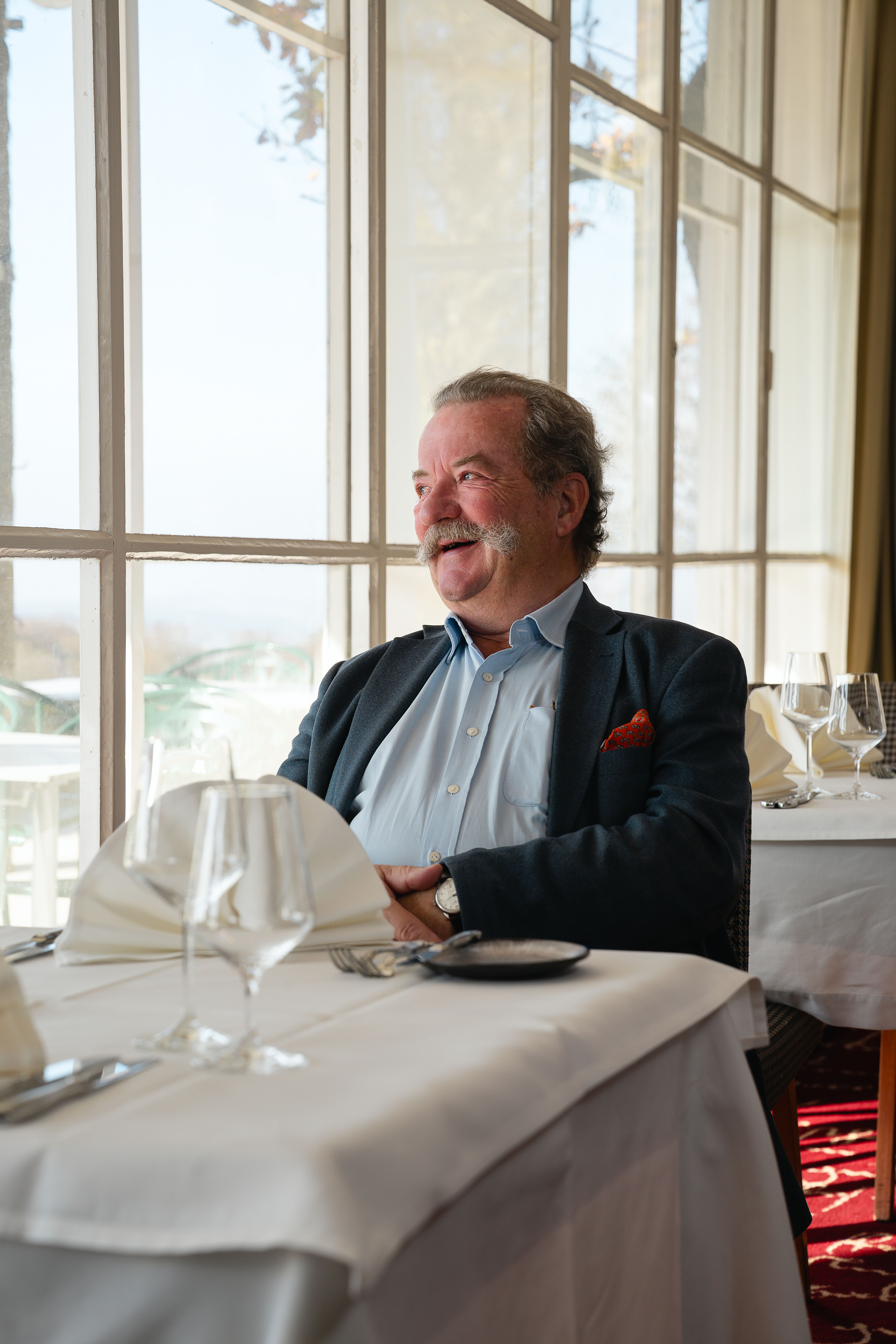 Frank Bläuel sitzt lächelnd im Restaurant des Berghotels Tulbingerkogel. Der Raum ist lichtdurchflutet, mit schön gedeckten Tischen, die der Umgebung eine warme und einladende Atmosphäre verleihen.