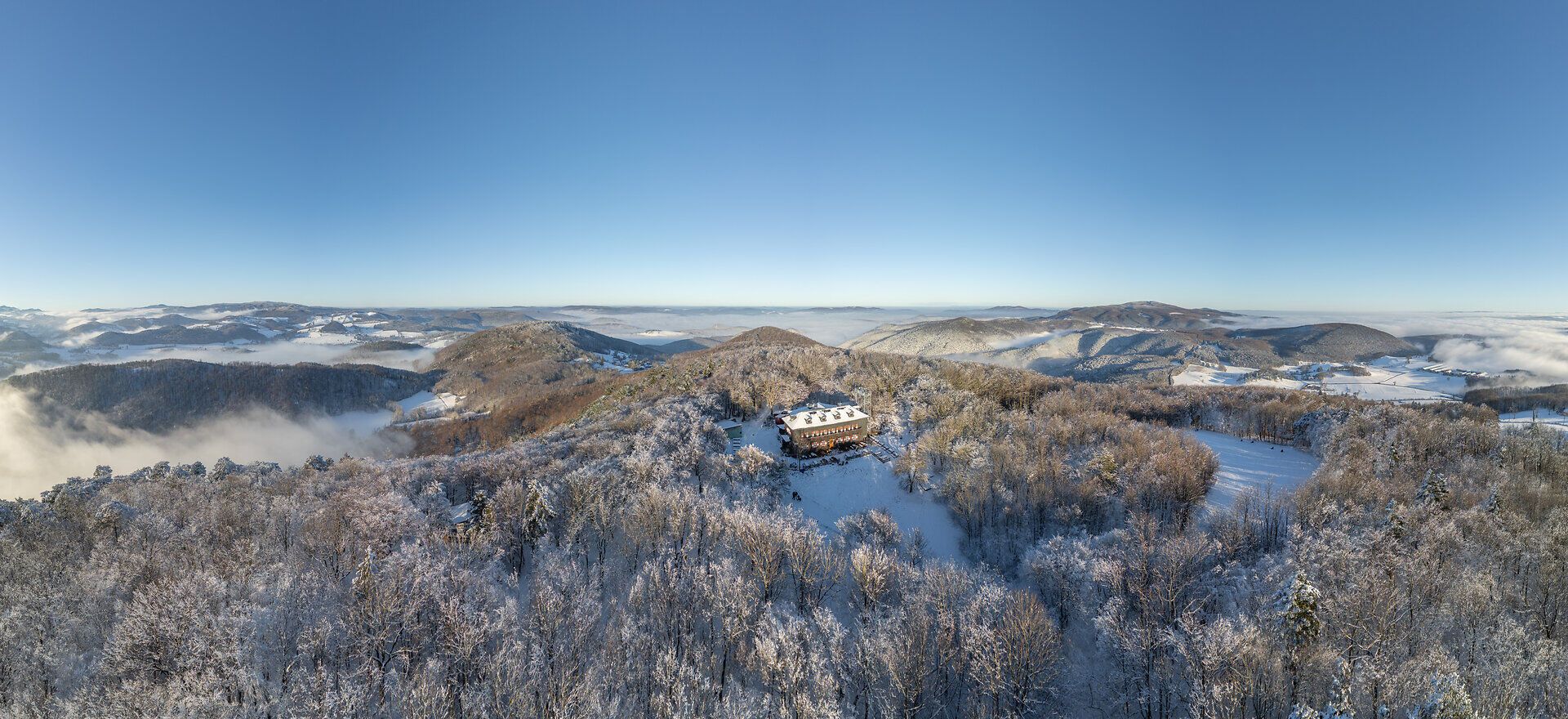 Luftaufnahme der verschneiten Peilsteinwände in Niederösterreich: Über den steilen, weiß überzogenen Kalkfelsen liegt klares Winterlicht, während das Schutzhaus am Gipfel in die Schneelandschaft eingebettet ist. Im Tal darunter ziehen dichte Nebelschwaden dahin, die sich vom sonnigen, strahlend blauen Himmel über dem Berg deutlich abheben.