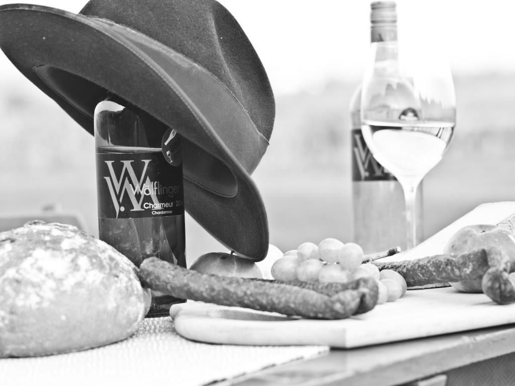 Schwarz-weiß Bild von Weinflaschen, Brot, Wurst und Trauben auf einem Tisch.