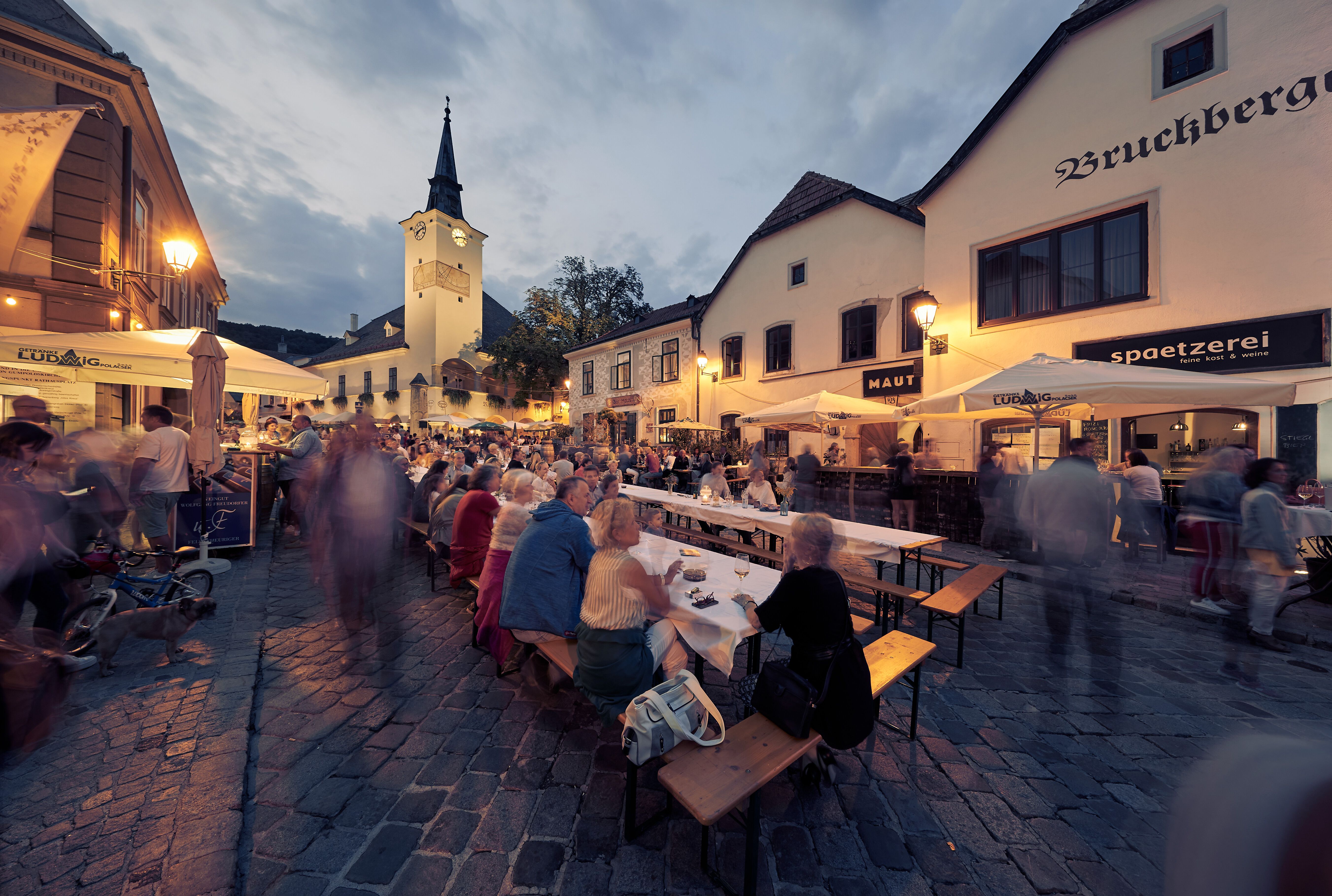 In der malerischen Kulisse des Wienerwaldes genießen die Gäste ein geselliges Beisammensein unter den schattigen Platanen. Der Duft von frisch gebackenem Brot und regionalen Weinen erfüllt die Luft und lädt zum Verweilen ein. Hier wird der Sommer in vollen Zügen gefeiert, während die sanften Hügel der Weinregion im Hintergrund leuchten.