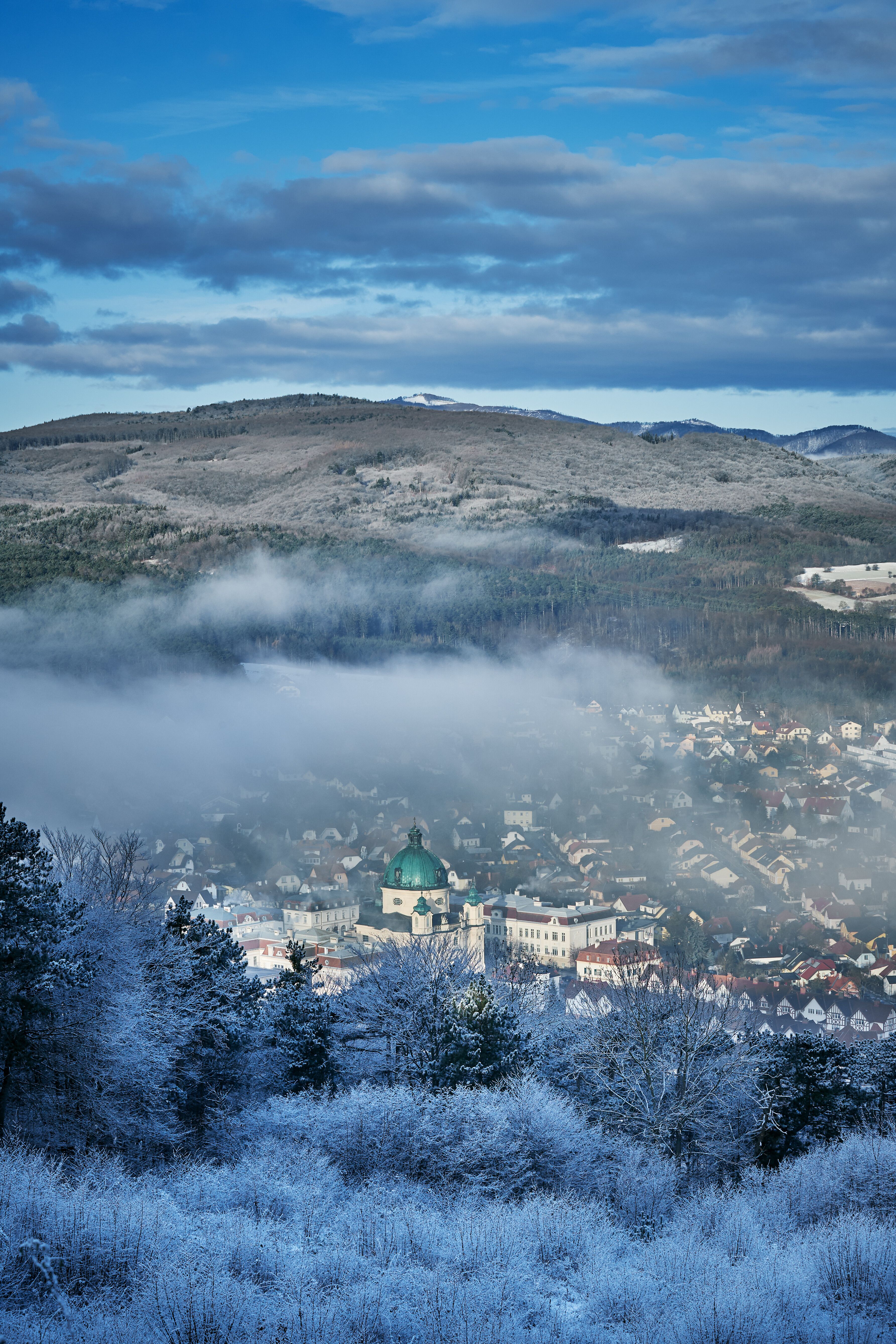 In der winterlichen Landschaft entfaltet sich eine zauberhafte Stille, während der Nebel sanft über die Dächer der Stadt gleitet. Die schneebedeckten Bäume und die sanften Hügel im Hintergrund schaffen eine malerische Kulisse, die zum Verweilen und Entdecken einlädt.