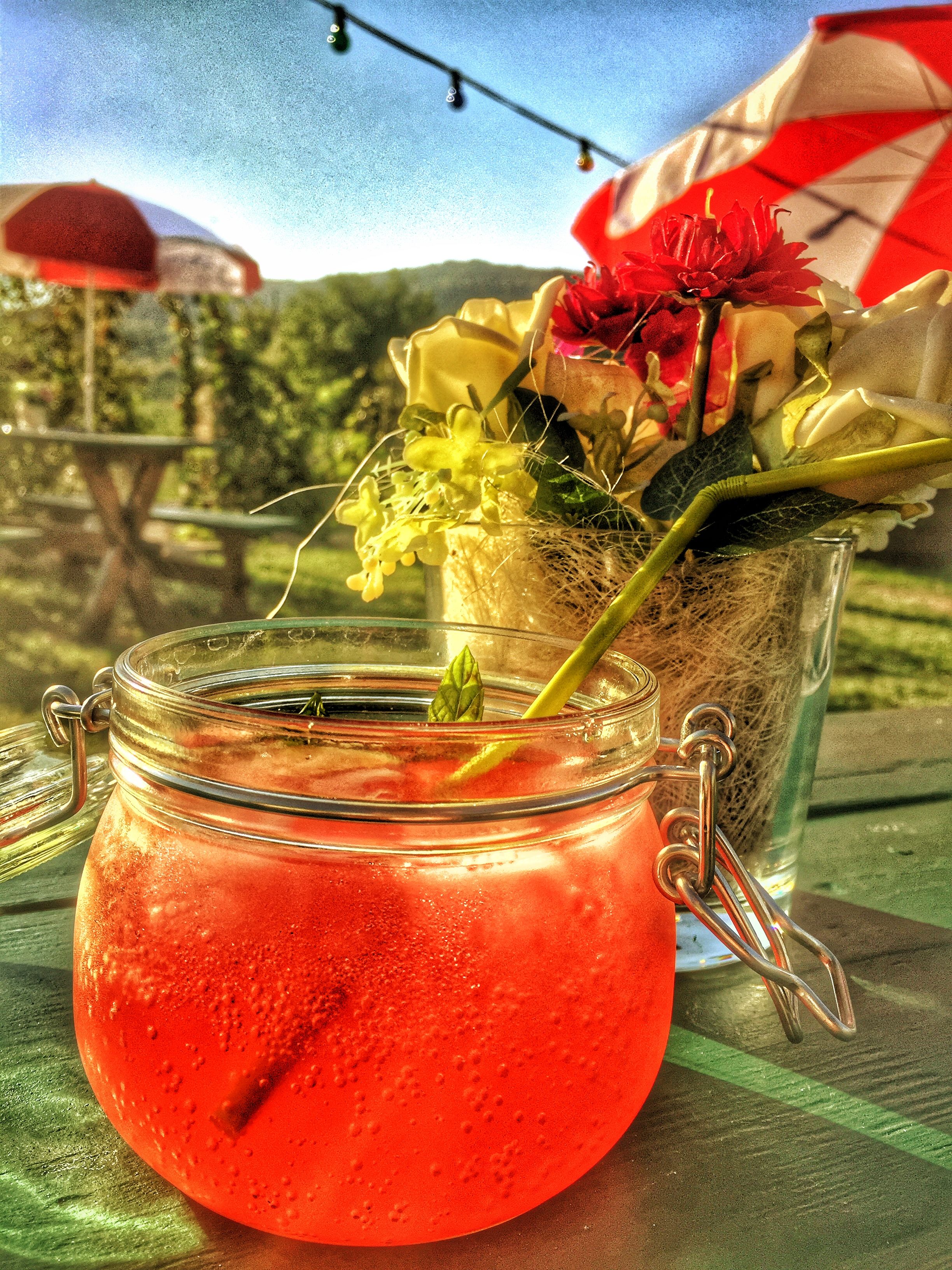 Ein rotes Getränk in einem Glas mit Bügelverschluss, daneben ein Blumenstrauß auf einem Tisch im Freien.