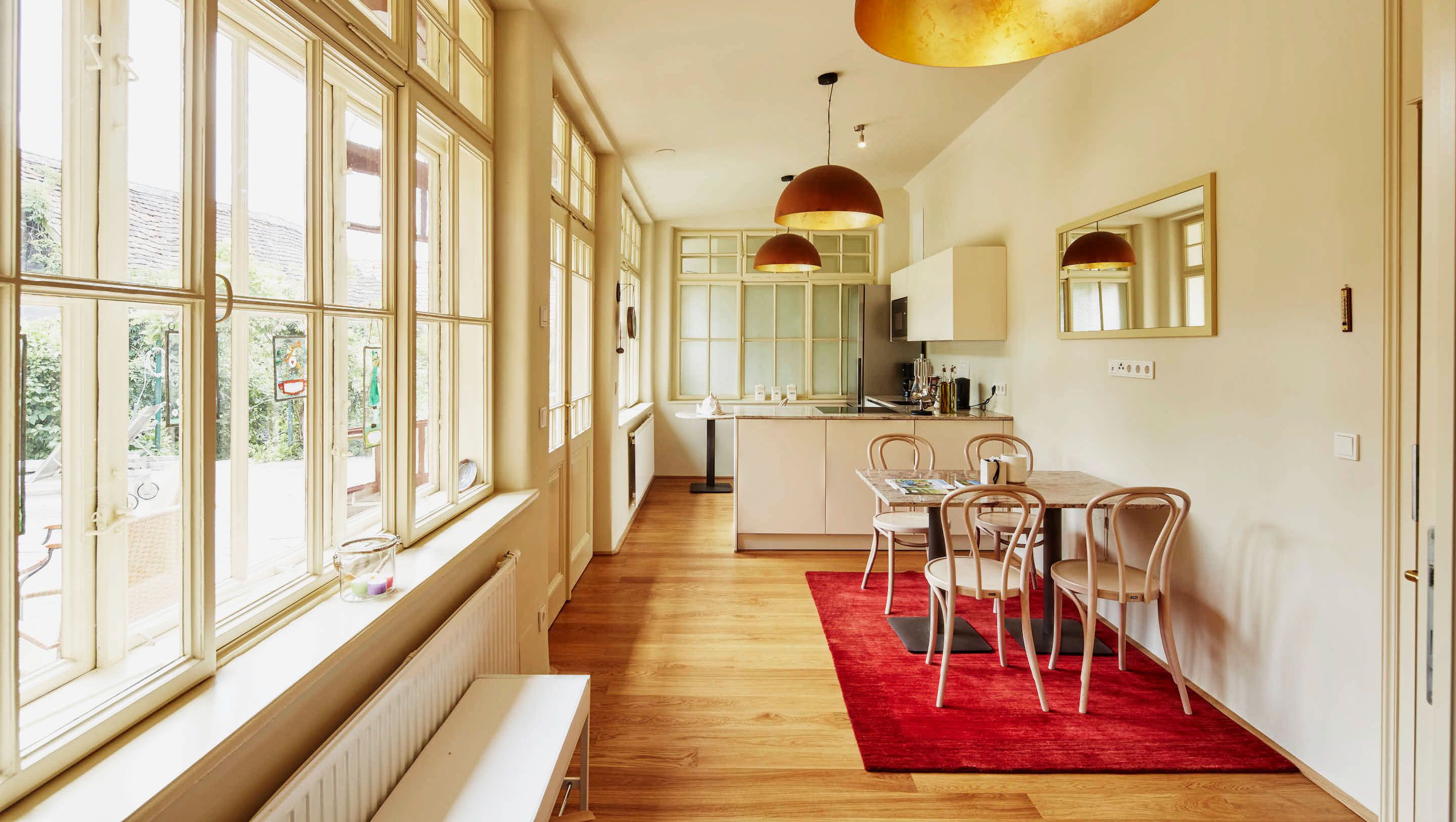 Helles Wohnzimmer mit Küche, Holzboden, roten Teppich, Esstisch und großen Fenstern.