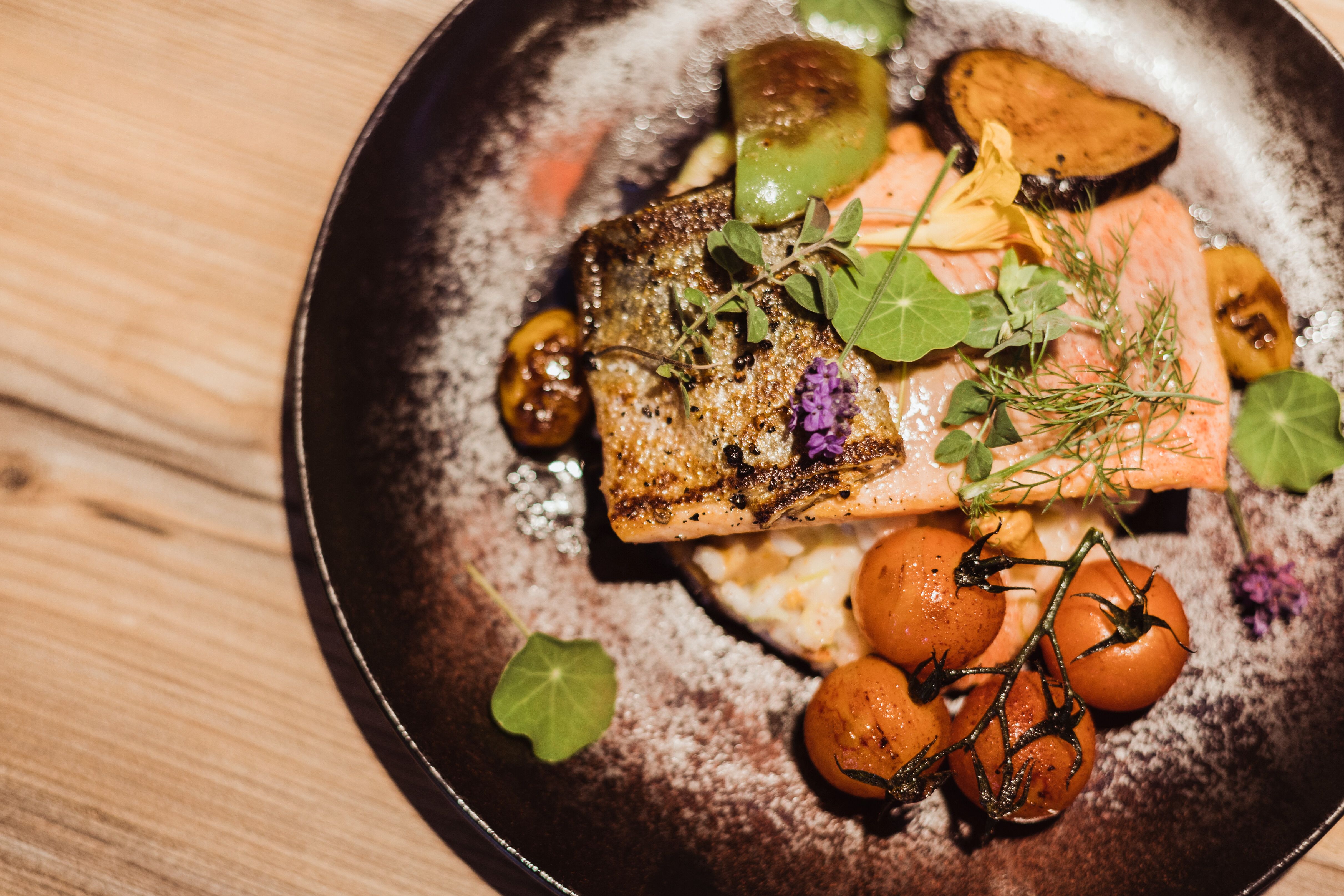 Ein köstliches Lachsforellenfilet, perfekt gebraten, liegt auf einem Bett aus cremigem Grammelrisotto, umgeben von buntem Gemüse und zarten Kräutern. Diese harmonische Komposition verspricht ein unvergessliches Geschmackserlebnis inmitten der idyllischen Kulisse des Wienerwaldes.