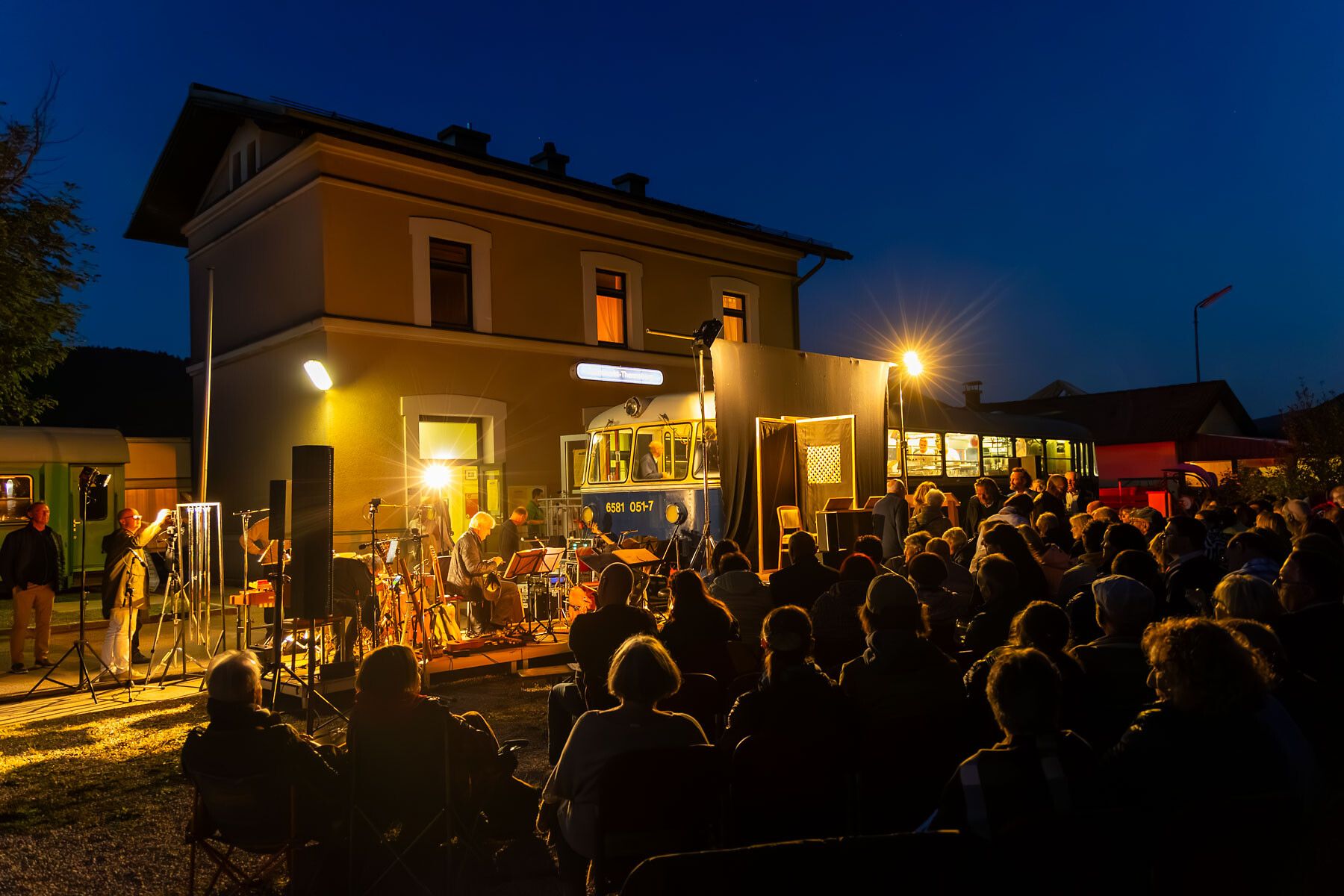 In der warmen Abenddämmerung versammeln sich Menschen vor dem Kulturbahnhof, um die Klänge eines Live-Konzerts zu genießen. Die Atmosphäre ist erfüllt von Musik und Lichtern, während die historische Kulisse des Bahnhofs eine besondere Stimmung schafft. Ein unvergesslicher Abend, der die Herzen der Besucher berührt.