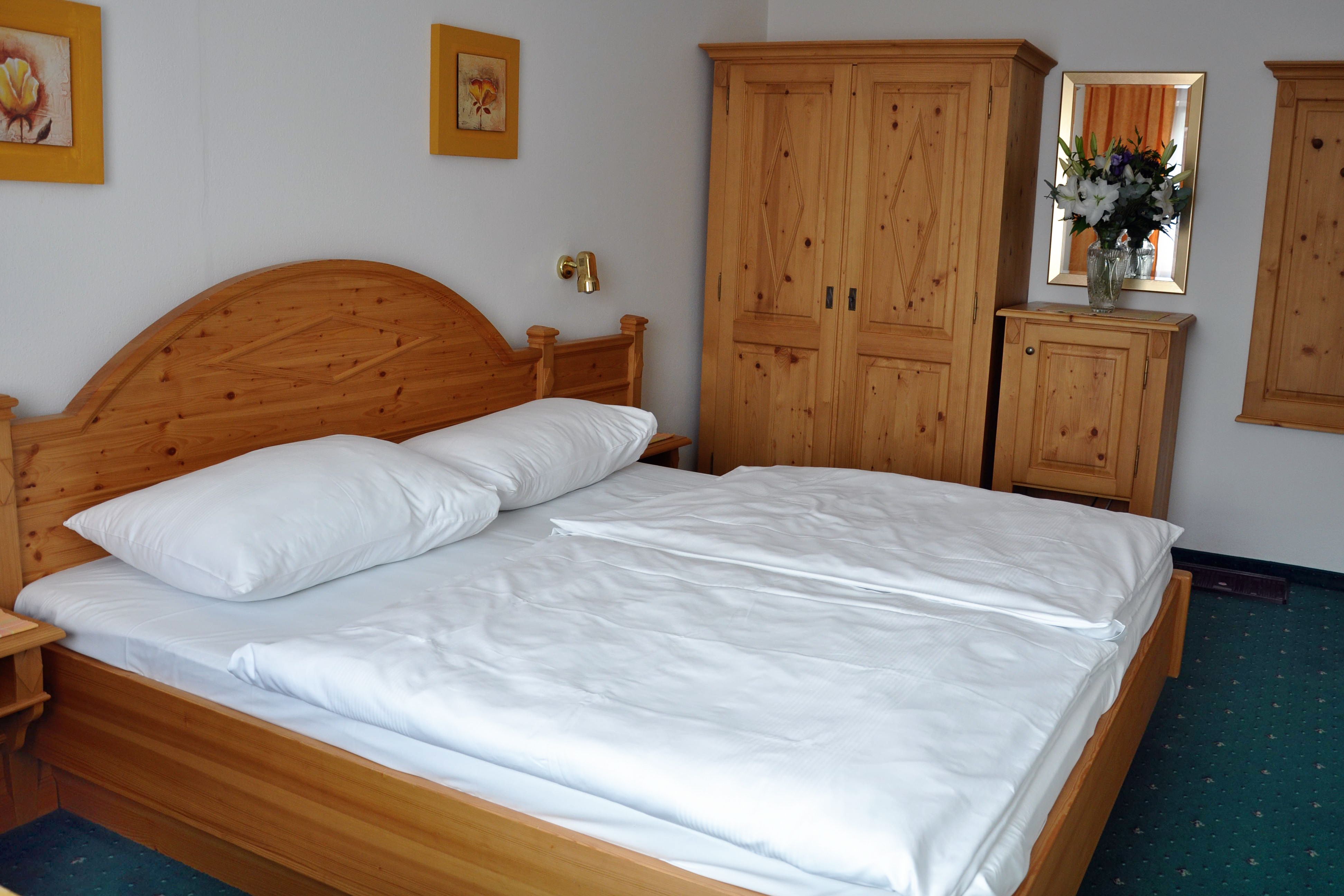 Komfortzimmer mit Vollholzmöbeln, Doppelbett, Kleiderschrank und Blumen auf Kommode.
