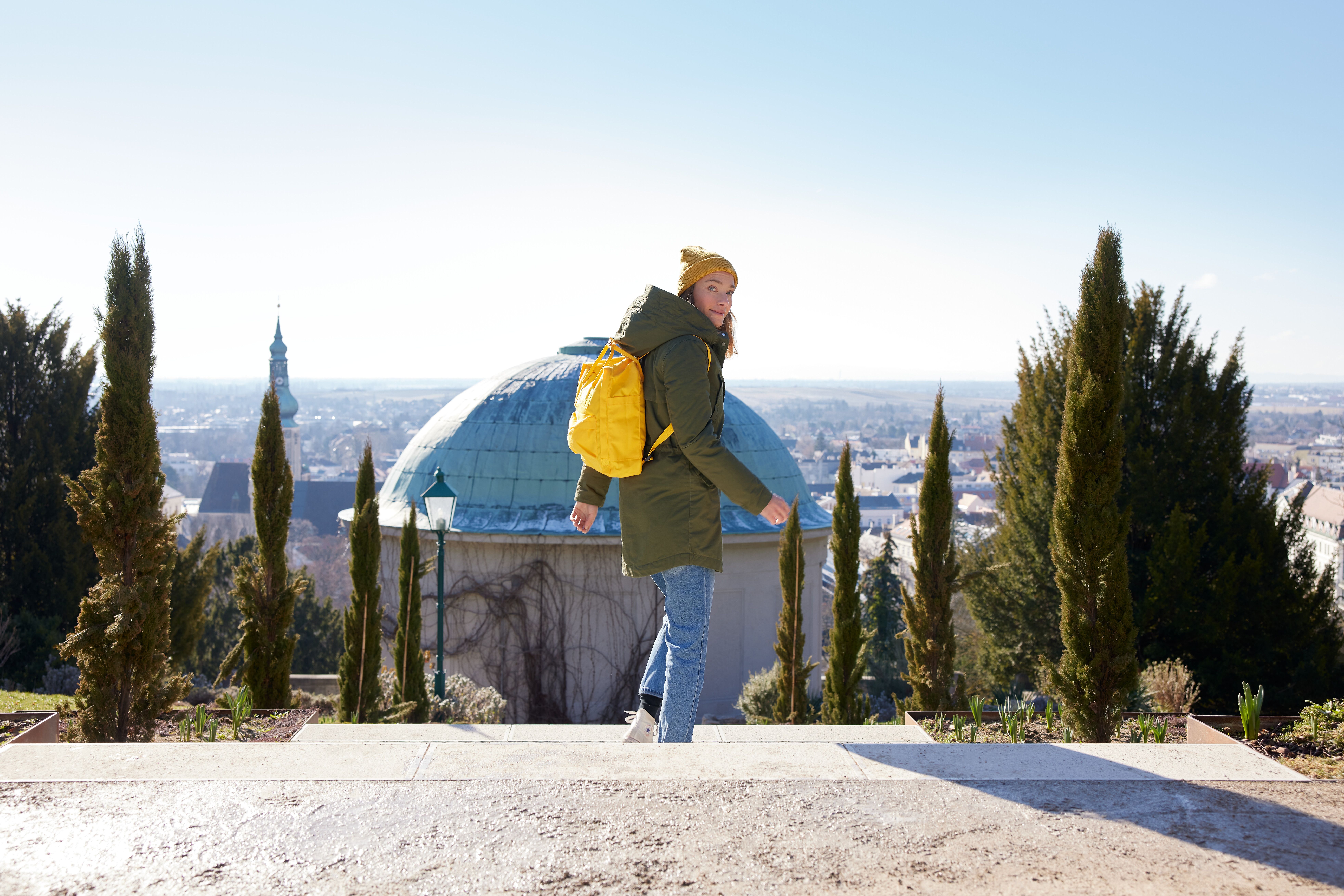 Ein strahlend blauer Himmel umrahmt die sanften Hügel, während eine Wanderin mit einem gelben Rucksack die malerische Aussicht genießt. Die frische Bergluft und die üppige Vegetation laden dazu ein, die Natur in vollen Zügen zu erleben und neue Energie zu tanken.
