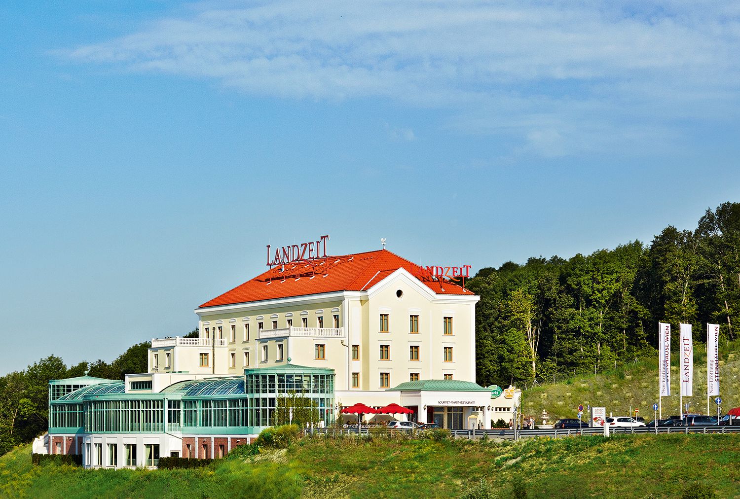 Landzeit Steinhäusl building with red roof and glass facade, surrounded by trees and blue sky.