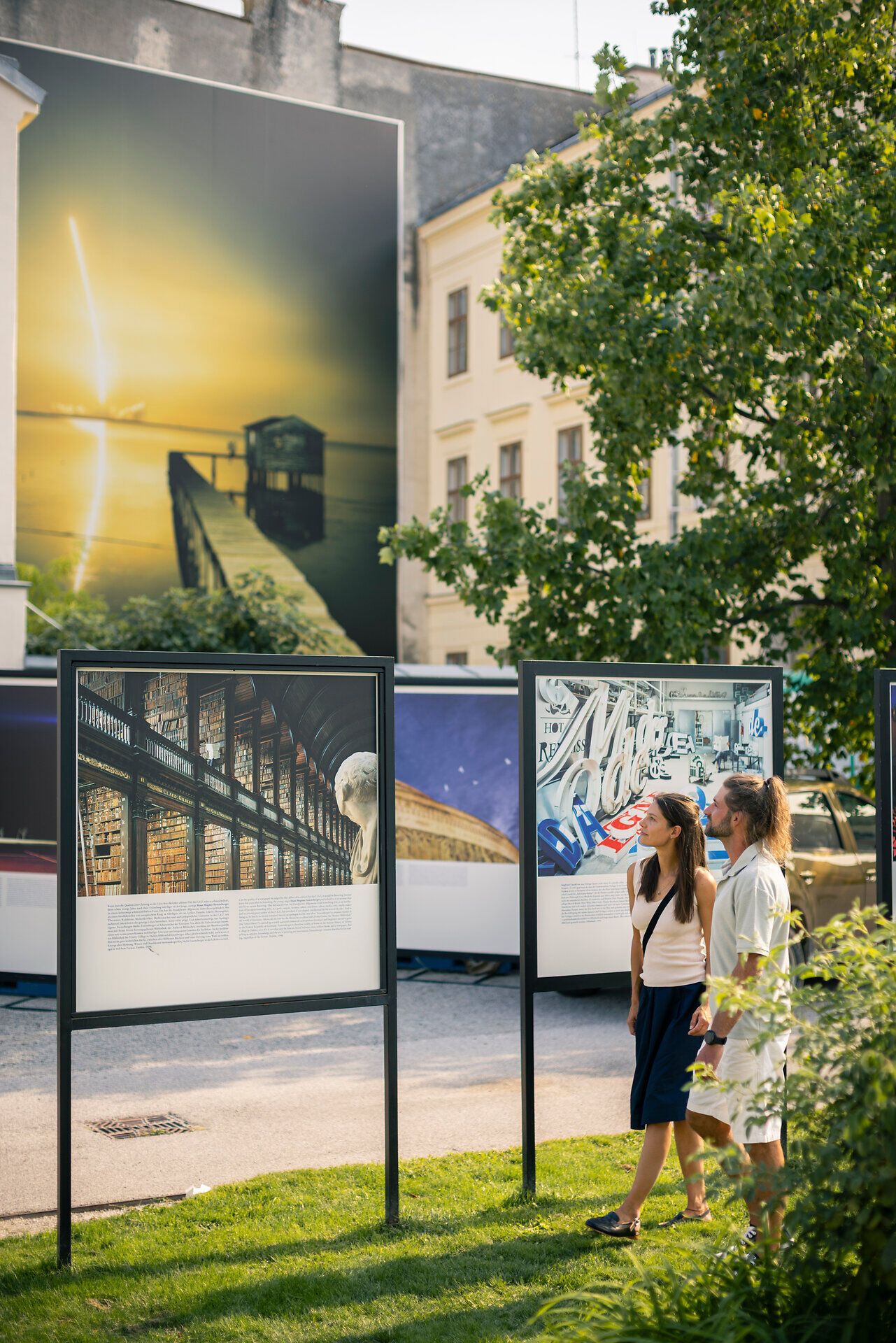 Eine Frau und ein Mann spazieren an einem warmen Sommertag in Baden bei Wien, während sie die Bilder des Fotofestivals La Gacilly Baden Photo begutachten.