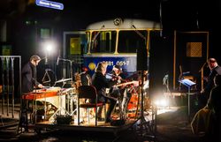 In der nächtlichen Kulisse des Kulturbahnhofs Altenmarkt entfaltet sich eine magische Atmosphäre, während Musiker harmonische Klänge erzeugen. Die sanften Lichter und die Präsenz der historischen Lokomotive schaffen einen einzigartigen Rahmen für ein unvergessliches Kulturereignis.