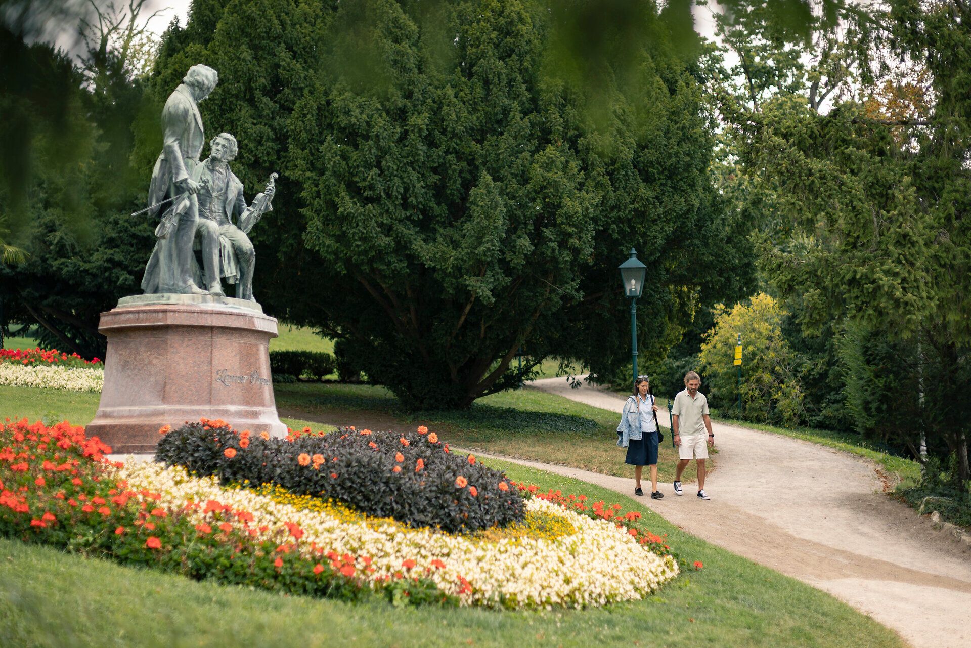 Eine Frau und ein Mann schlendern im Kurpark Baden am Lanner Strauß Denkmal vorbei. Vor ihnen erstreckt sich ein großes Blumenbeet mit roten, gelben und weißen Blumen. Im Hintergrund sind Bäume des Parks sichtbar.