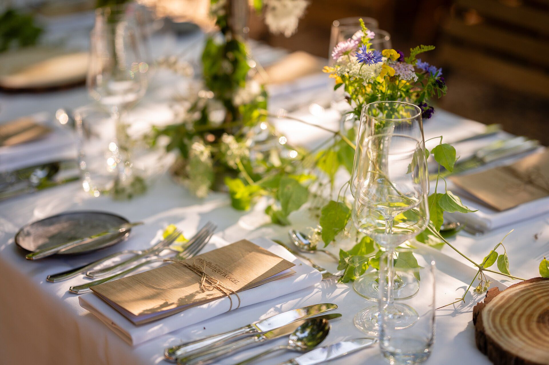 Ein elegant gedeckter Tisch steht inmitten eines Waldes, das weiße Tischtuch kontrastiert mit der natürlichen Umgebung. Grüne Blumenarrangements zieren den Tisch, während die Sonnenstrahlen durch das Blätterdach fallen und ihn in warmes Licht tauchen. Weingläser warten auf die Gäste und versprechen ein genussvolles Erlebnis unter freiem Himmel.