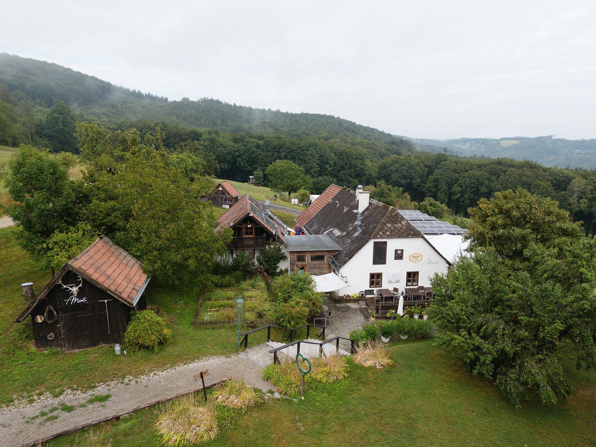 Das Gasthaus Mirli, eingebettet im malerischen Irenental, aus der Vogelperspektive betrachtet. Umgeben von den dichten, grünen Wäldern des Wienerwalds, zieht eine mystische Atmosphäre auf, die das Bild in einen geheimnisvollen, bezaubernden Zauber hüllt.