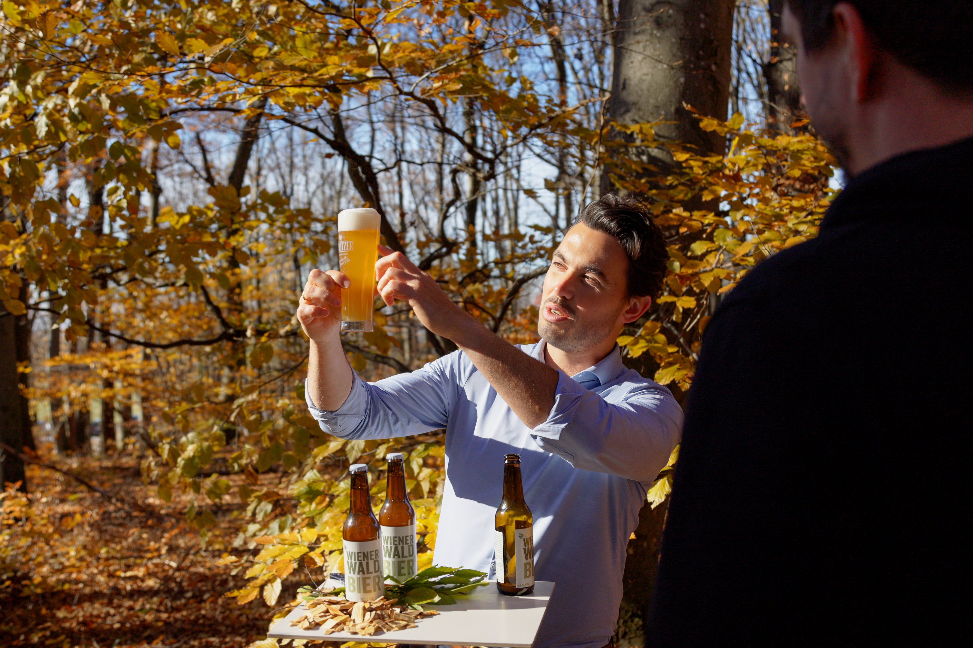 Im herbstlichen Wienerwald bei strahlendem Sonnenschein und gelblich orange verfärbten Blättern wird das Wienerwaldbier, eingeschenkt in einem Bierglas, von dem Guide in das Sonnenlicht gehalten.