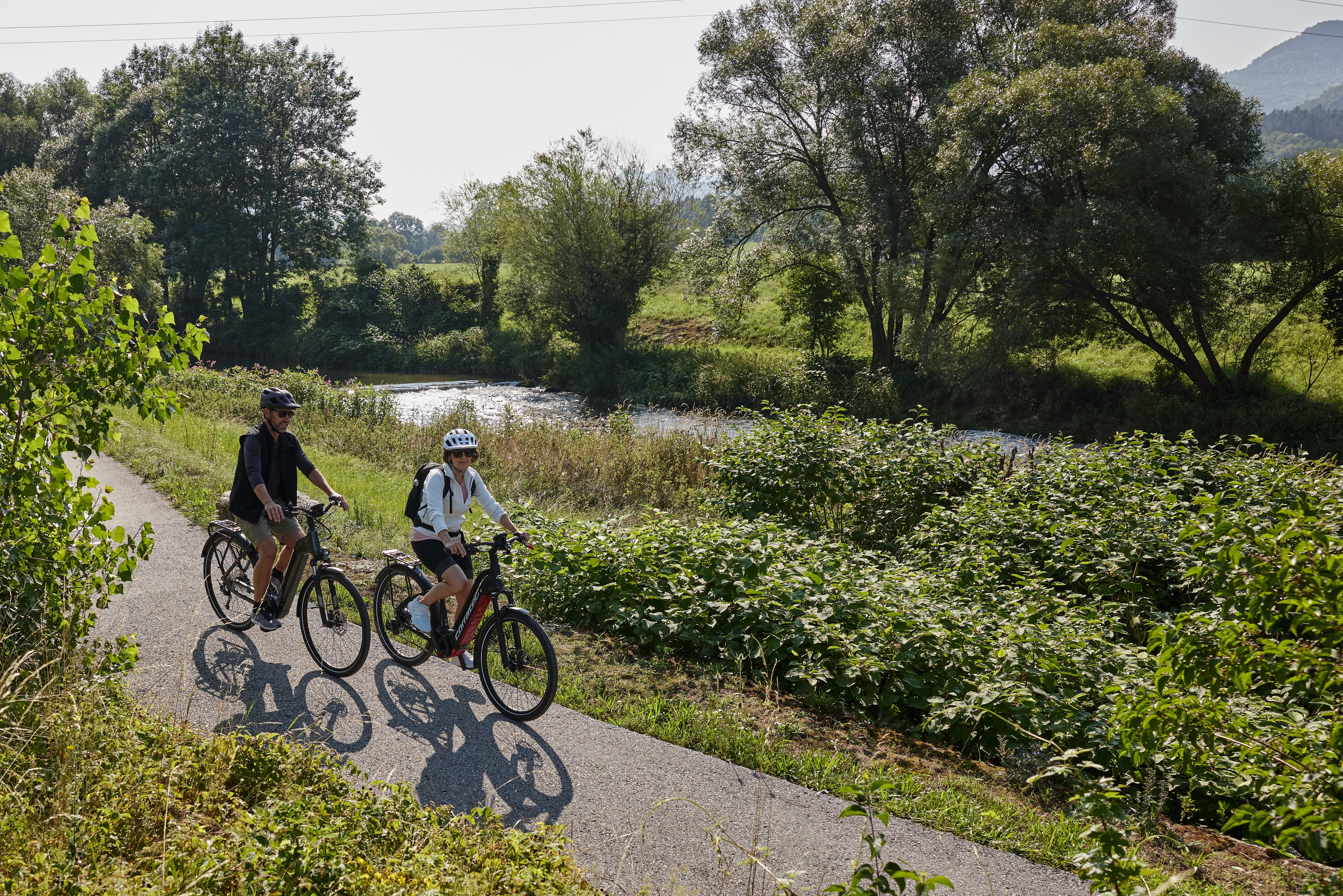 Radfahrer gleiten entspannt entlang des malerischen Radwegs, umgeben von üppigem Grün und sanften Hügeln. Die ruhige Atmosphäre und das Plätschern des nahen Baches laden dazu ein, die Schönheit der Natur zu genießen und neue Energie zu tanken.