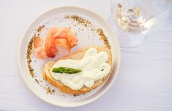 Weiß gedeckter Tisch mit weißem Teller in freundlicher Atmosphäre; darauf Baguette mit heller Creme, grünem Spargel und Prosciutto, daneben ein Glas Weißwein.