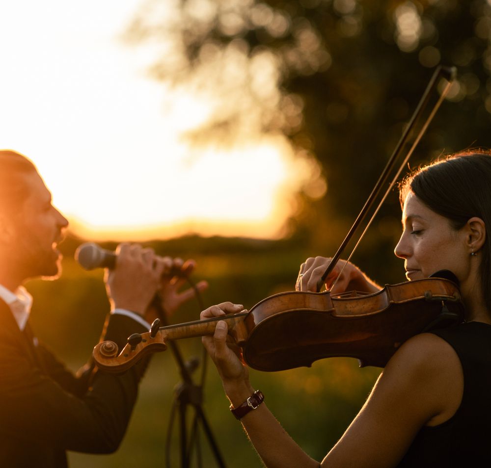 Live-Musik bei Sonnenuntergang im Weingut Herzog in Bad Vöslau, mit Sänger am Mikrofon und Geigerin im Vordergrund, eingebettet in stimmungsvolle Weinlandschaft.