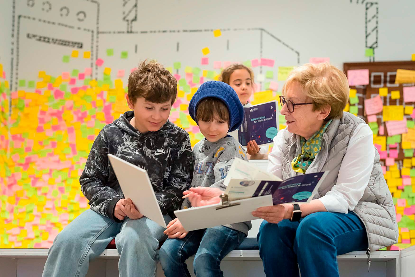 Eine ältere Frau und zwei Kinder sitzen zusammen und lesen in einem Raum mit bunten Haftnotizen an der Wand.