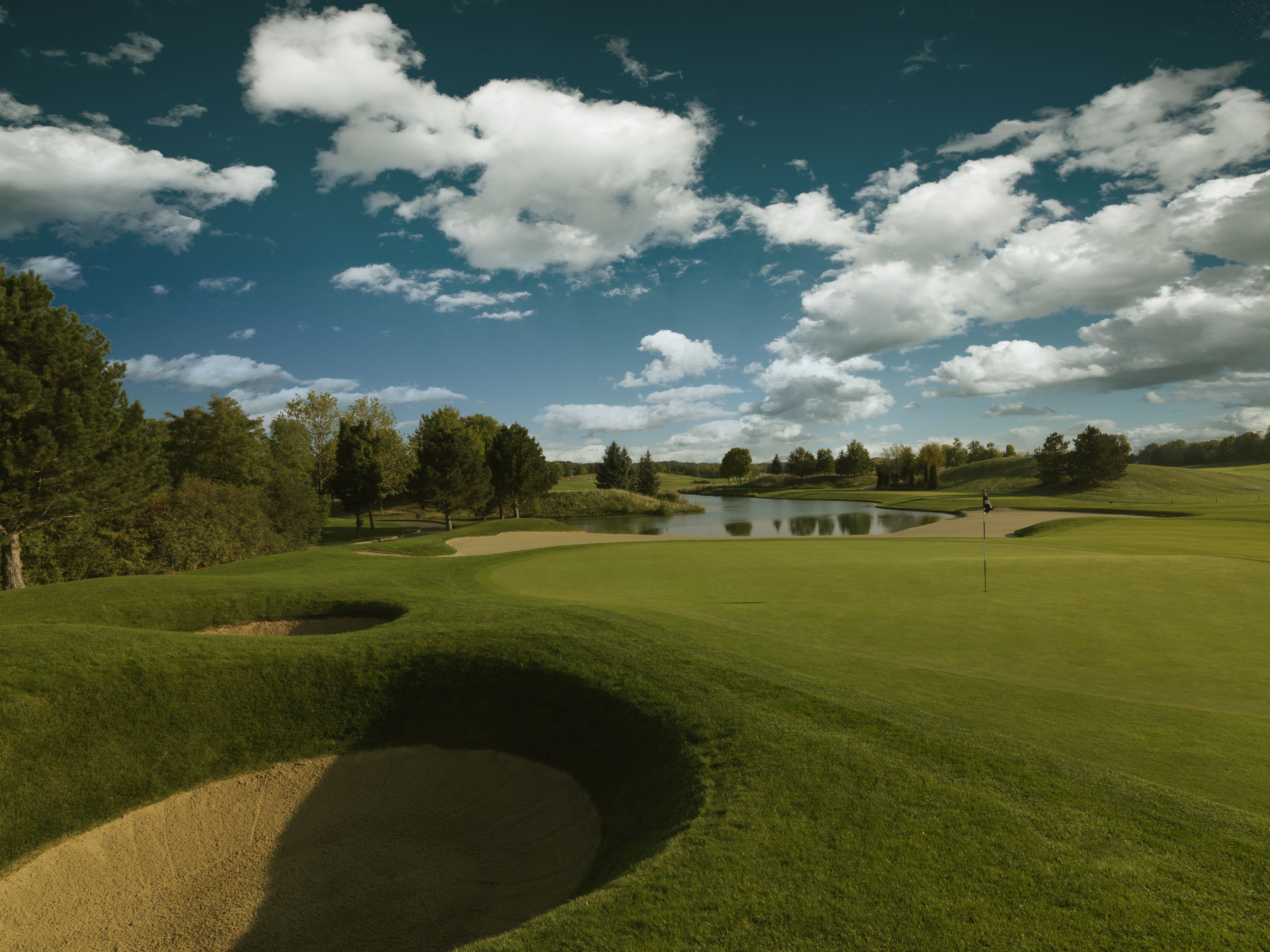 Golfplatz mit Sandbunker, Grün und Teich unter blauem Himmel mit Wolken.